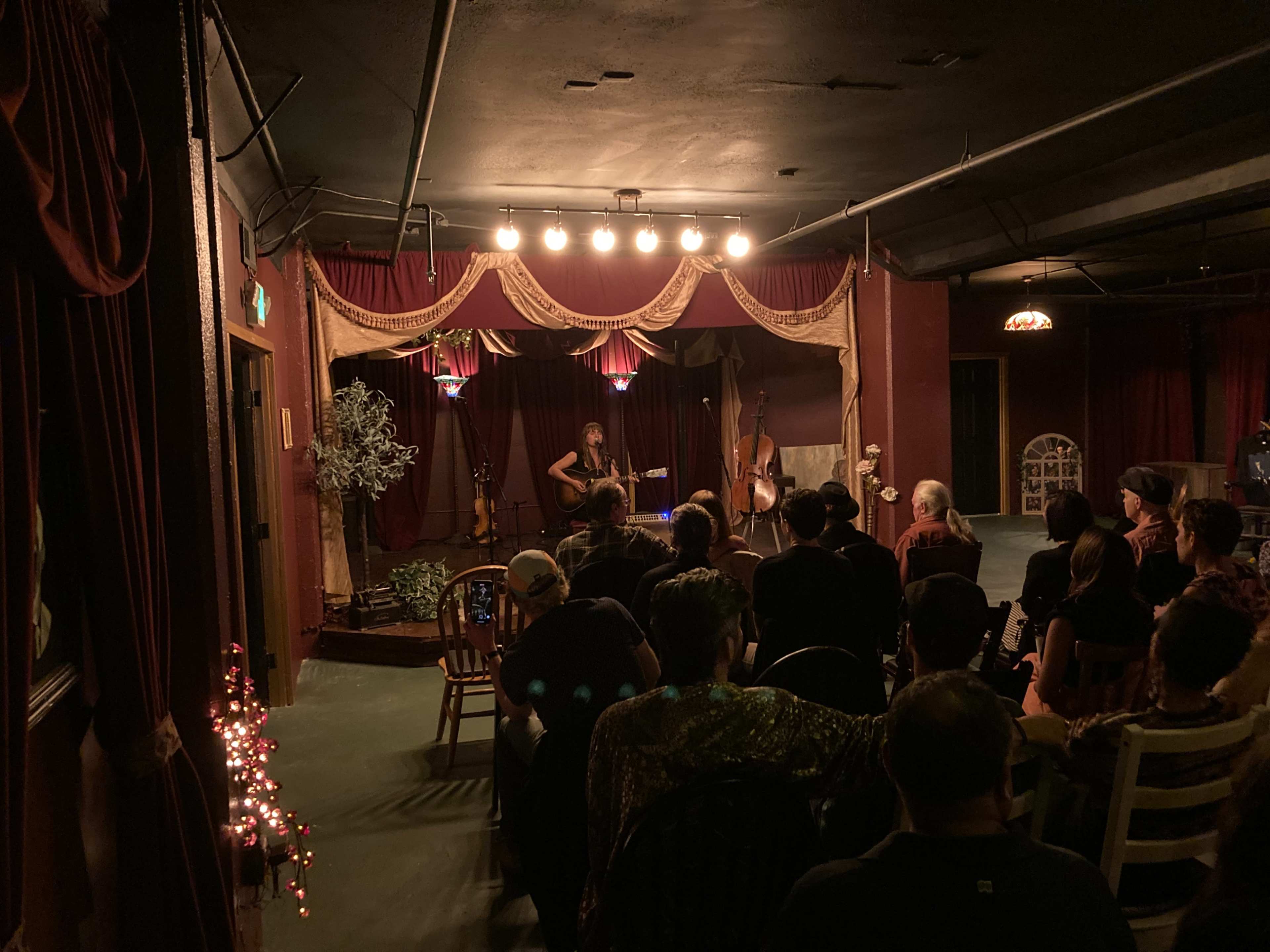 A musician performs on stage in a dimly lit venue while an audience watches from their seats.