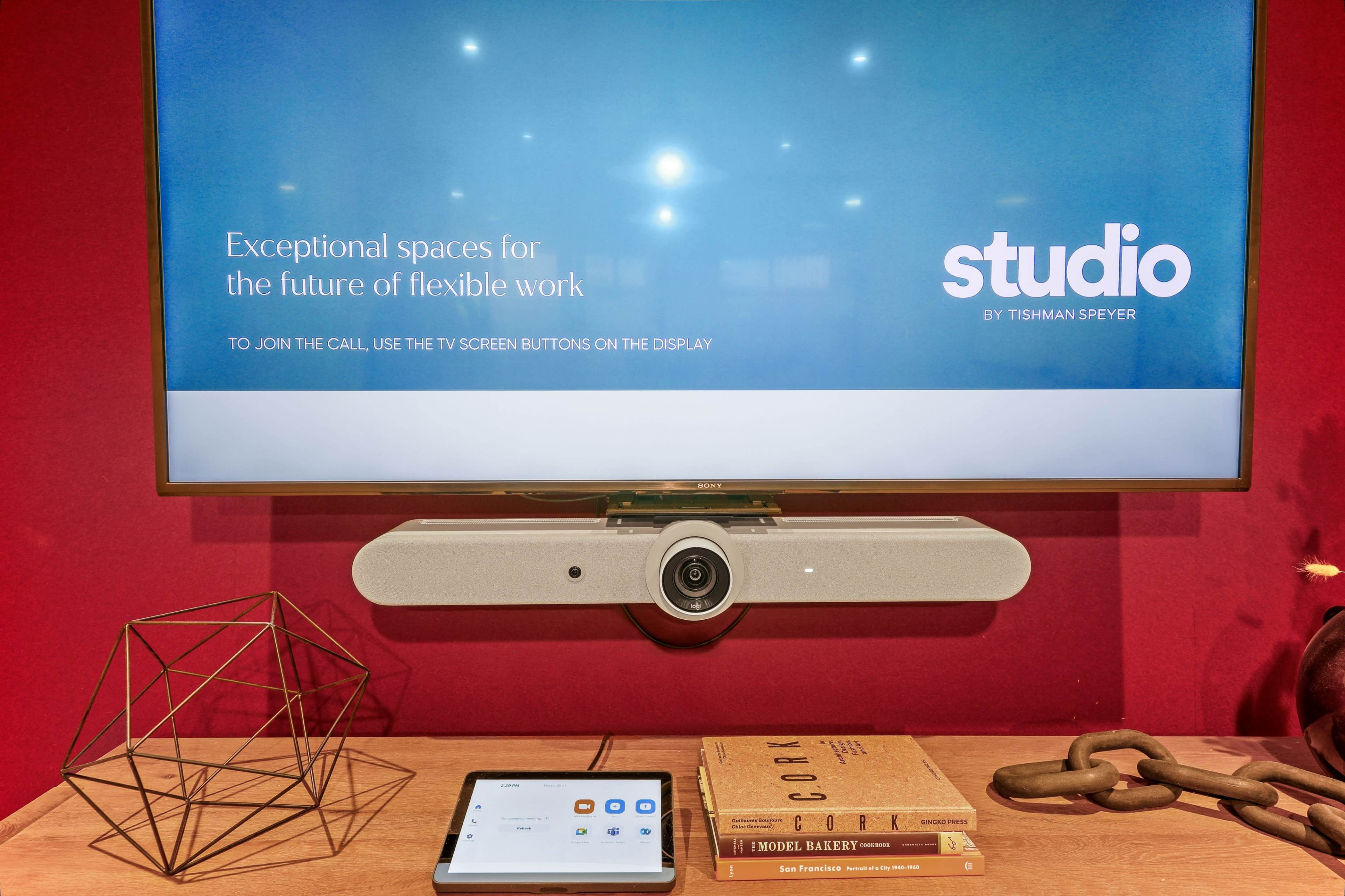 A television mounted on a red wall displays a presentation about flexible work, accompanied by a tablet and decorative objects on a wooden table.