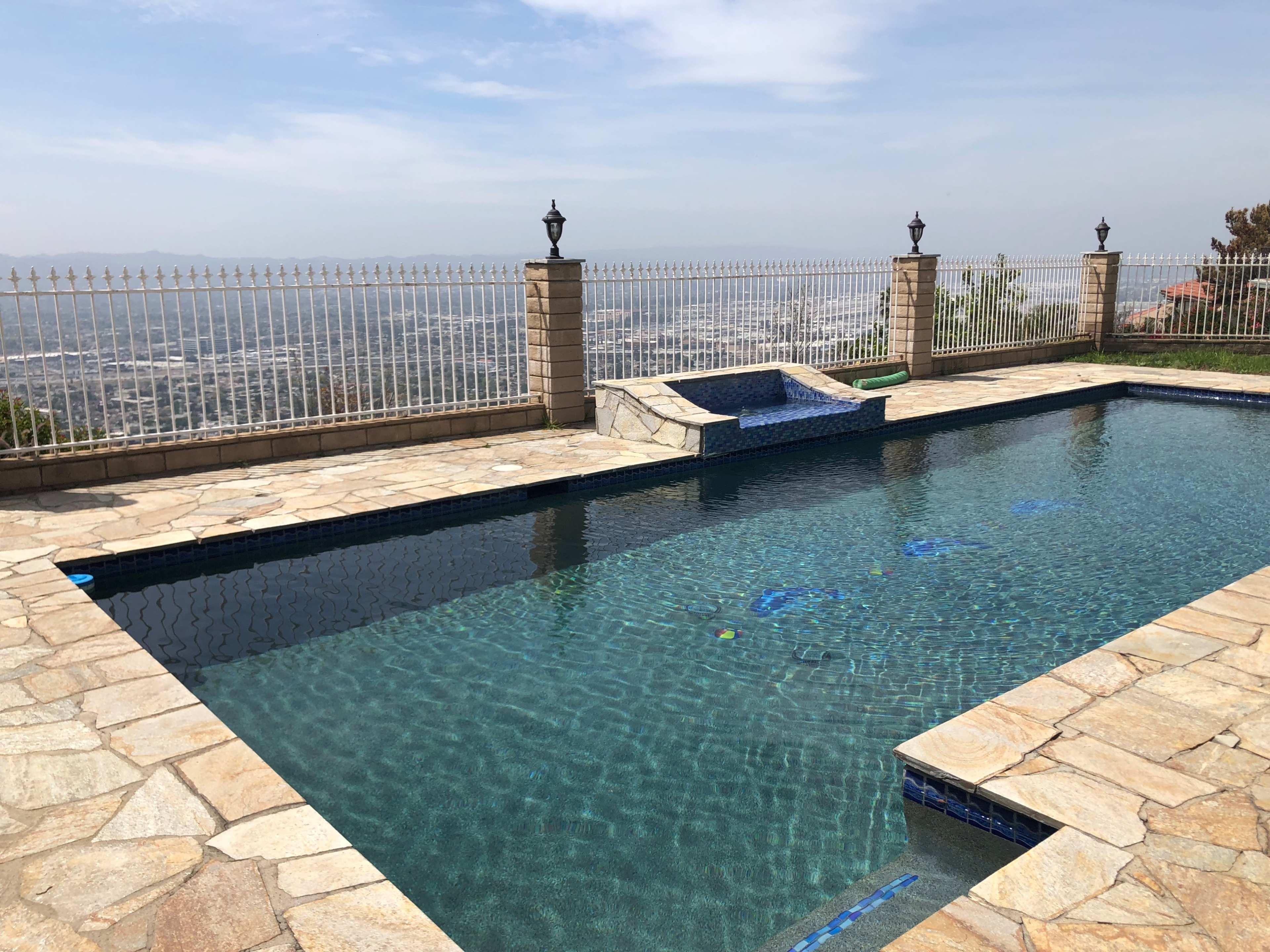 The image shows a swimming pool with a stone deck overlooking a distant landscape, enclosed by a wrought iron fence.