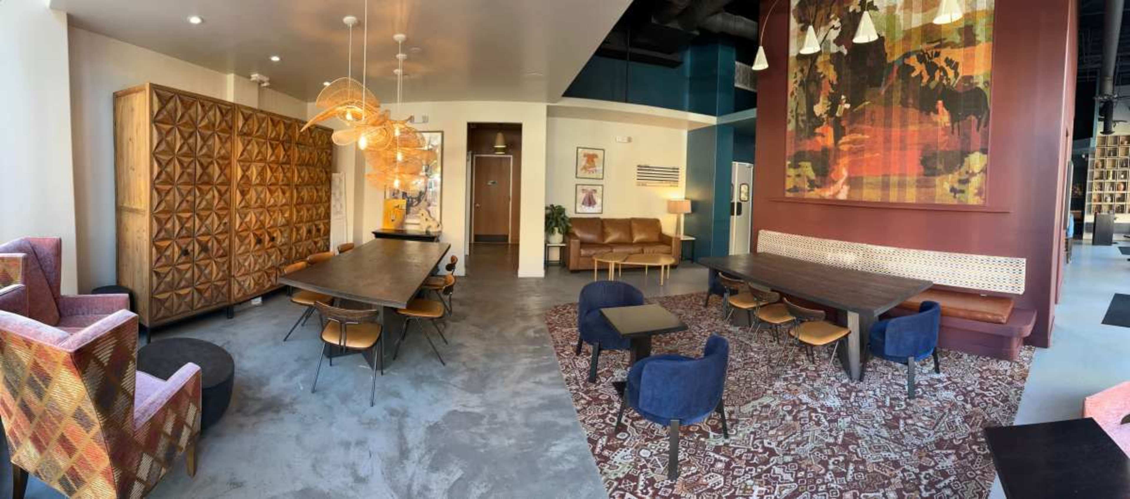 The image shows a contemporary interior space featuring various seating arrangements, decorative lighting, and a large patterned wall art piece.