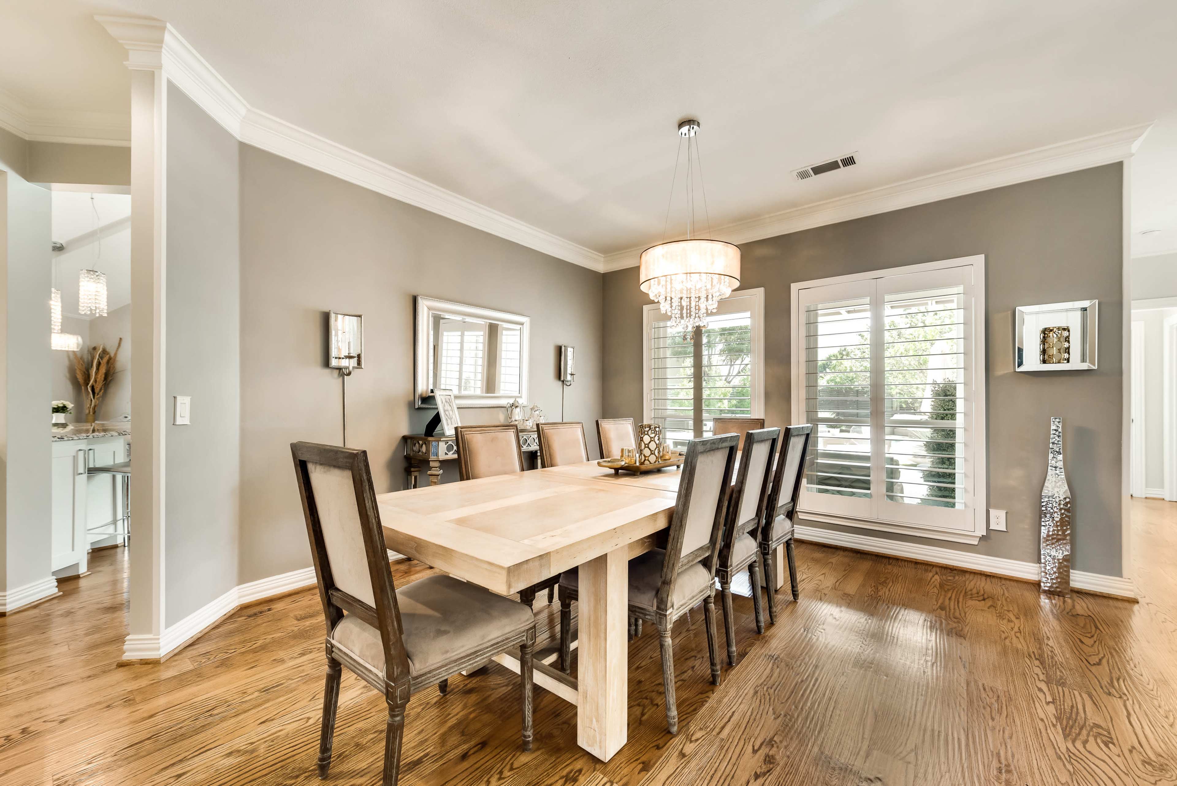 A well-lit dining room features a wooden table surrounded by eight upholstered chairs, with large windows displaying outdoor greenery.