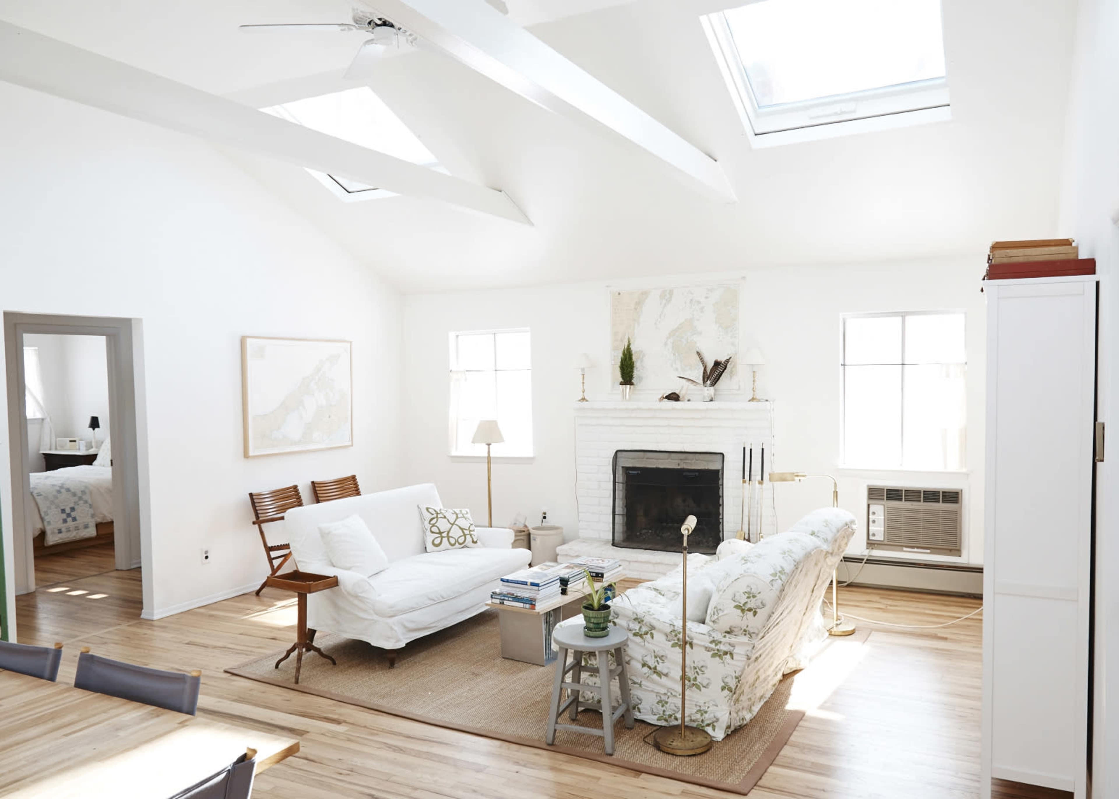 A bright, airy living room featuring a white sofa, beige armchair, and a fireplace, with large windows and skylights illuminating the space.