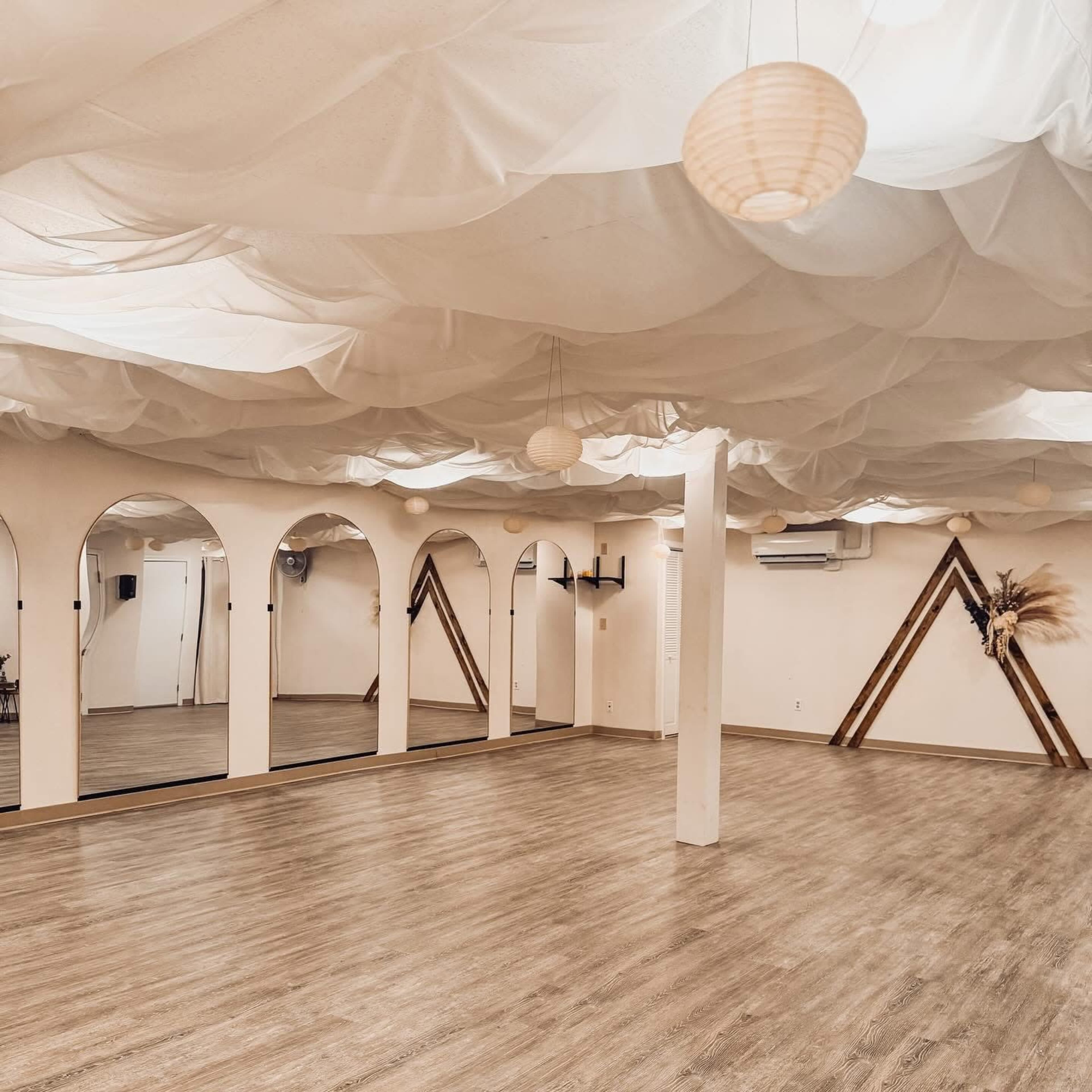 The image shows a spacious practice studio with a textured wooden floor, large mirrors on one wall, and a ceiling draped with white fabric.