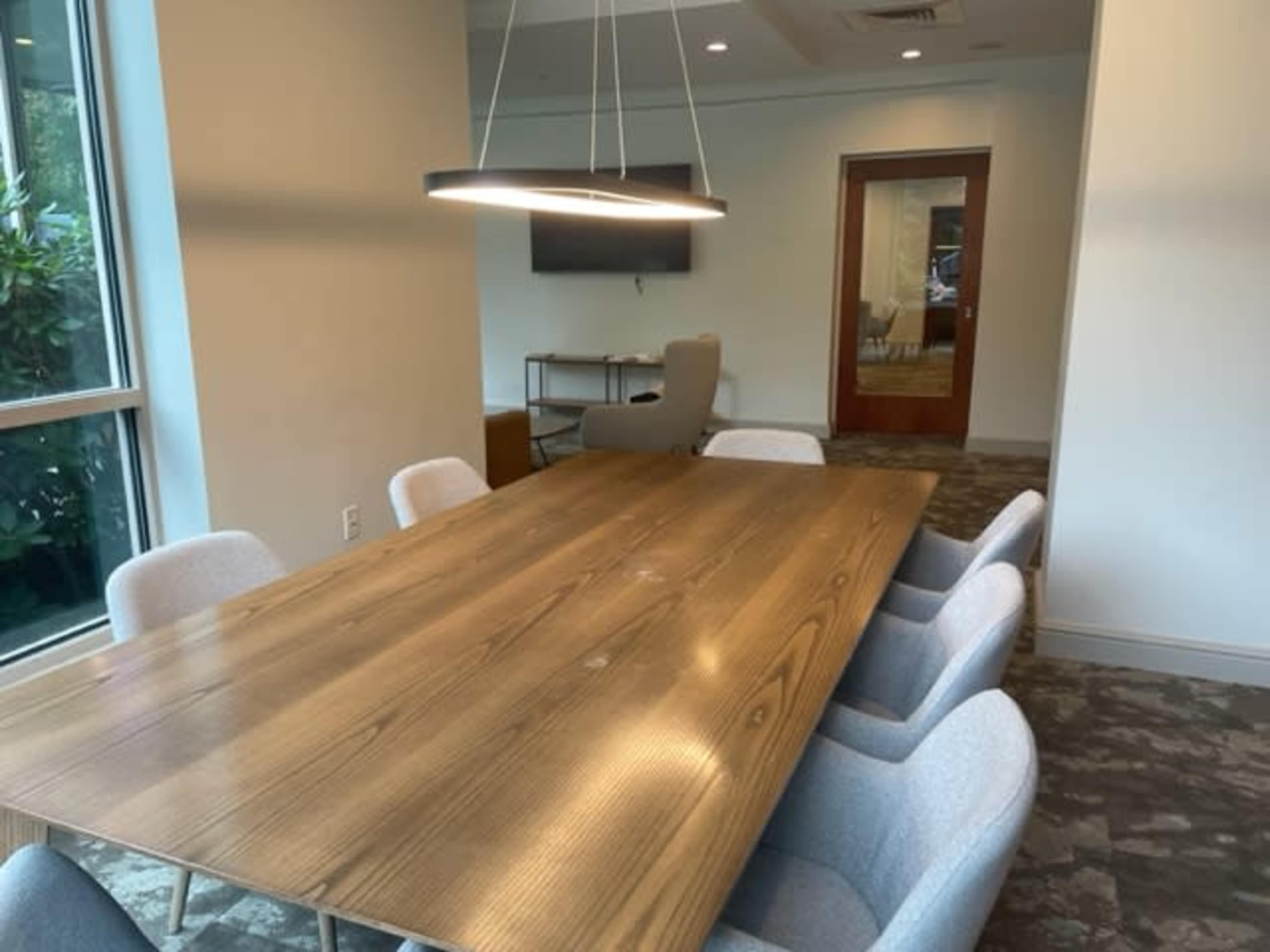 A conference room with a large wooden table surrounded by several light-colored chairs, and a wall-mounted screen in the background.