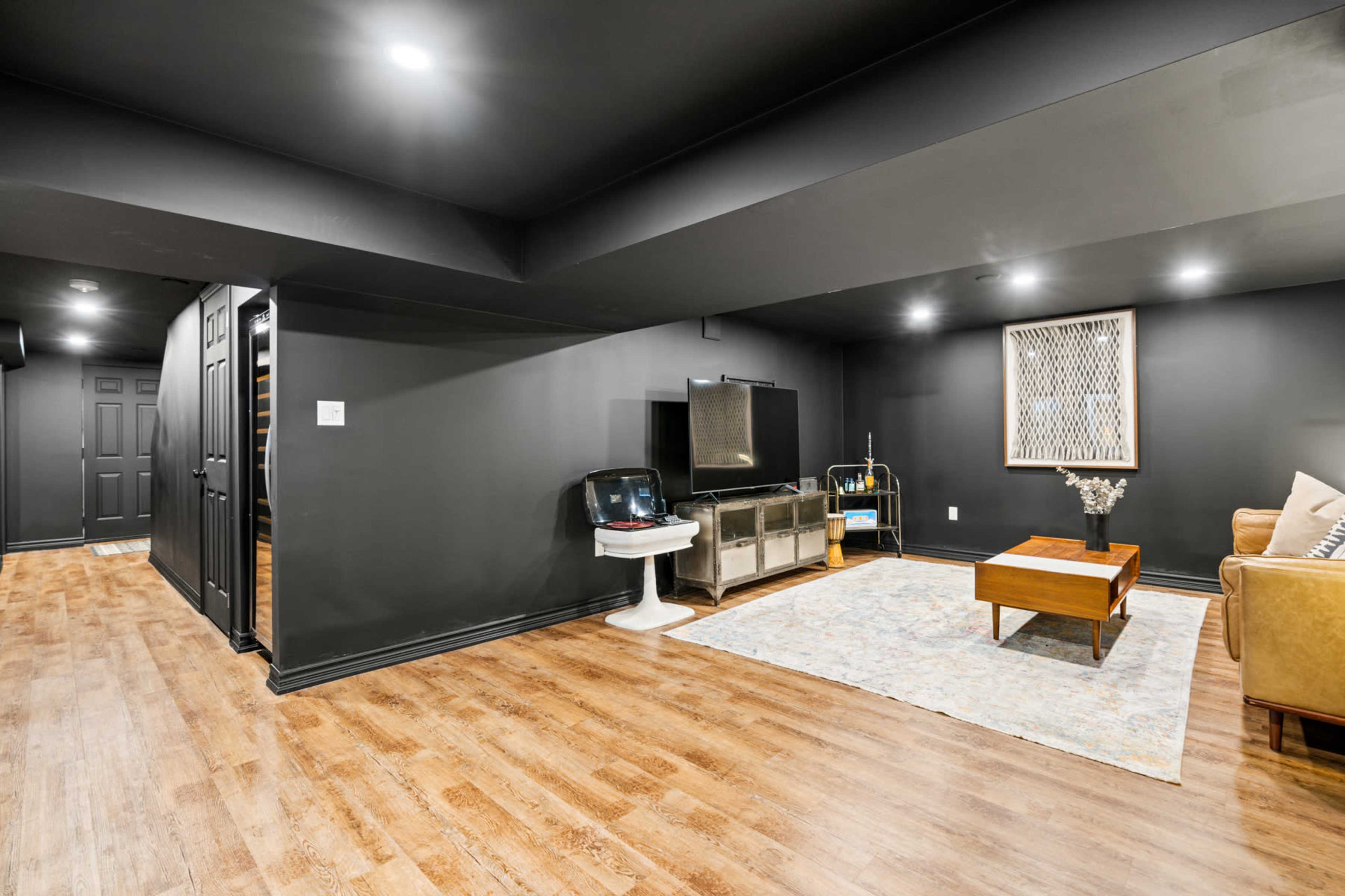 The scene shows a finished basement with hardwood floors, dark walls, a coffee table, a couch, and a television mounted on a stand.