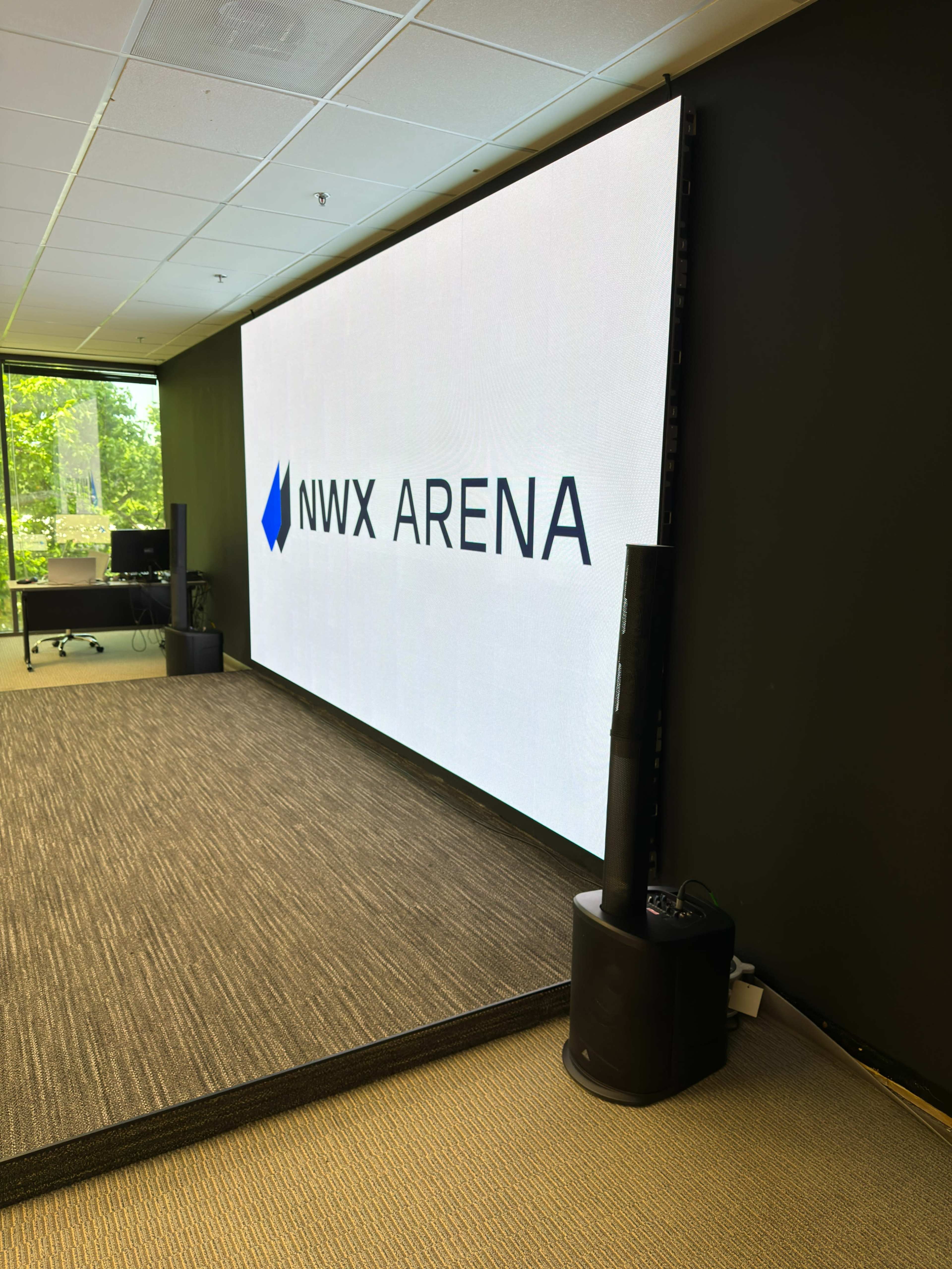The image shows a large digital screen displaying the text "NWX ARENA" in a modern office space with green outdoor views visible through the windows.