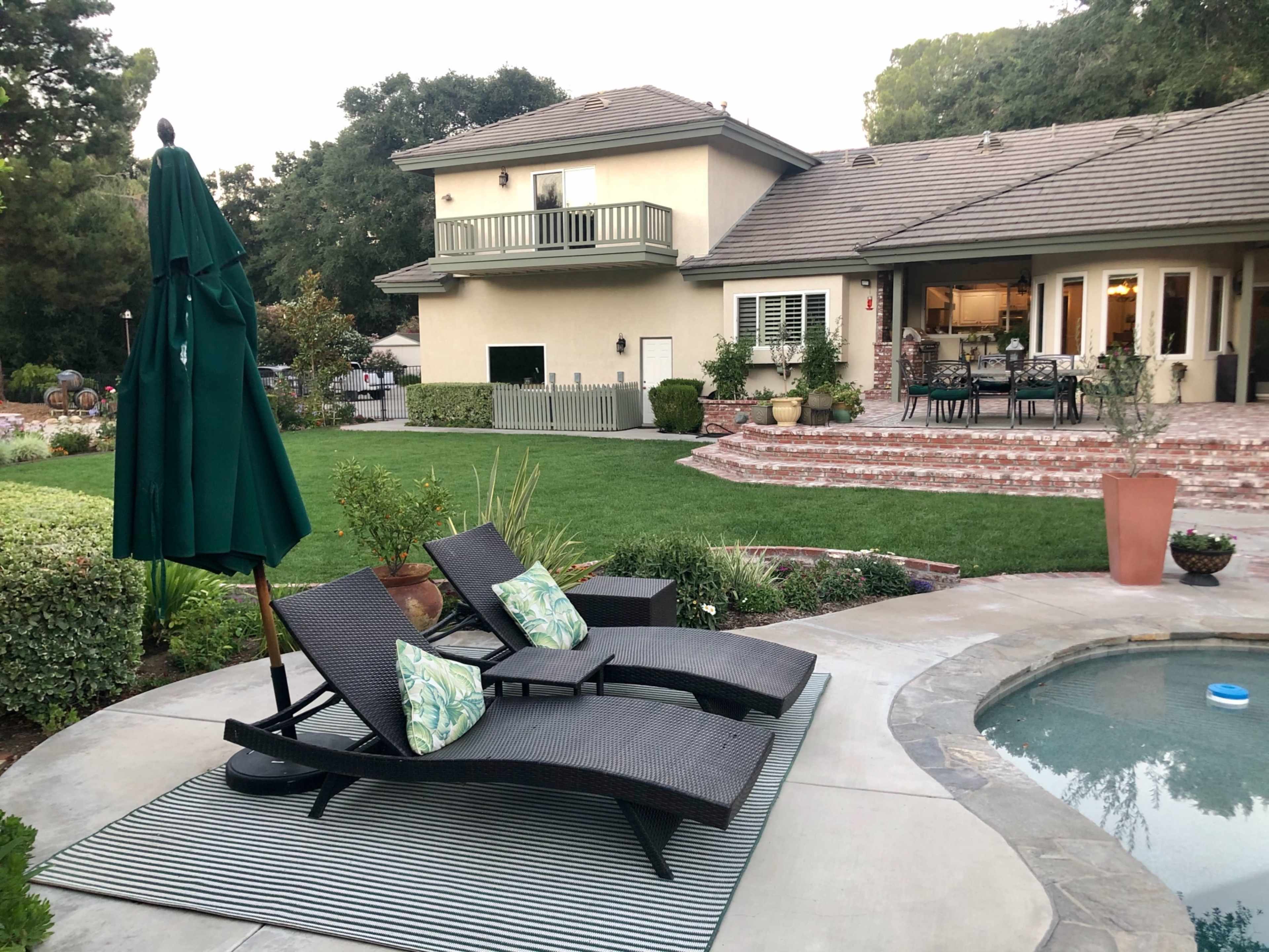 A pair of lounge chairs with cushions sits on a striped rug near a pool in a landscaped backyard behind a two-story house.