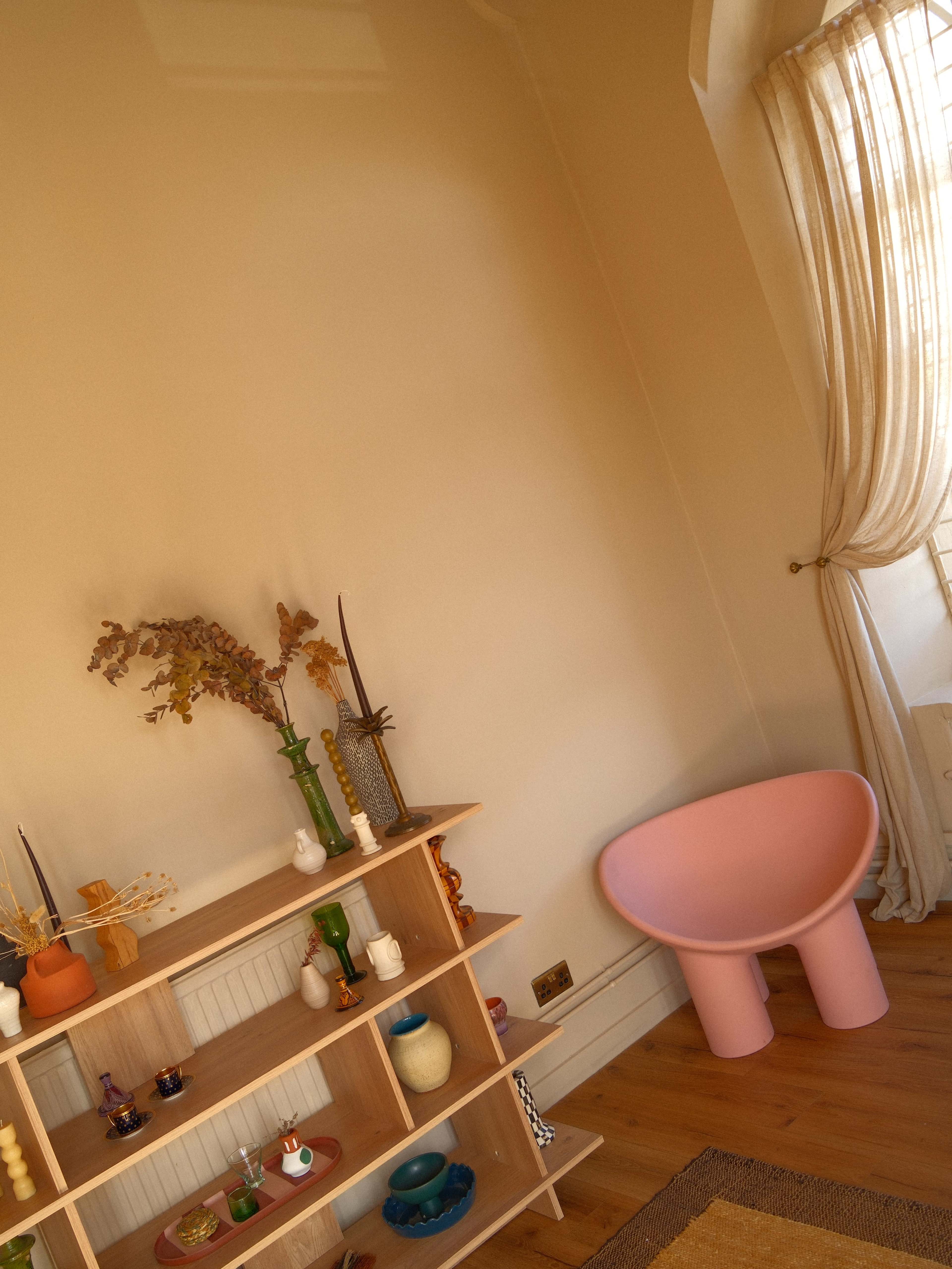 The image shows a corner of a room featuring a wooden shelf adorned with decorative objects and a pink sculptural chair beside it.