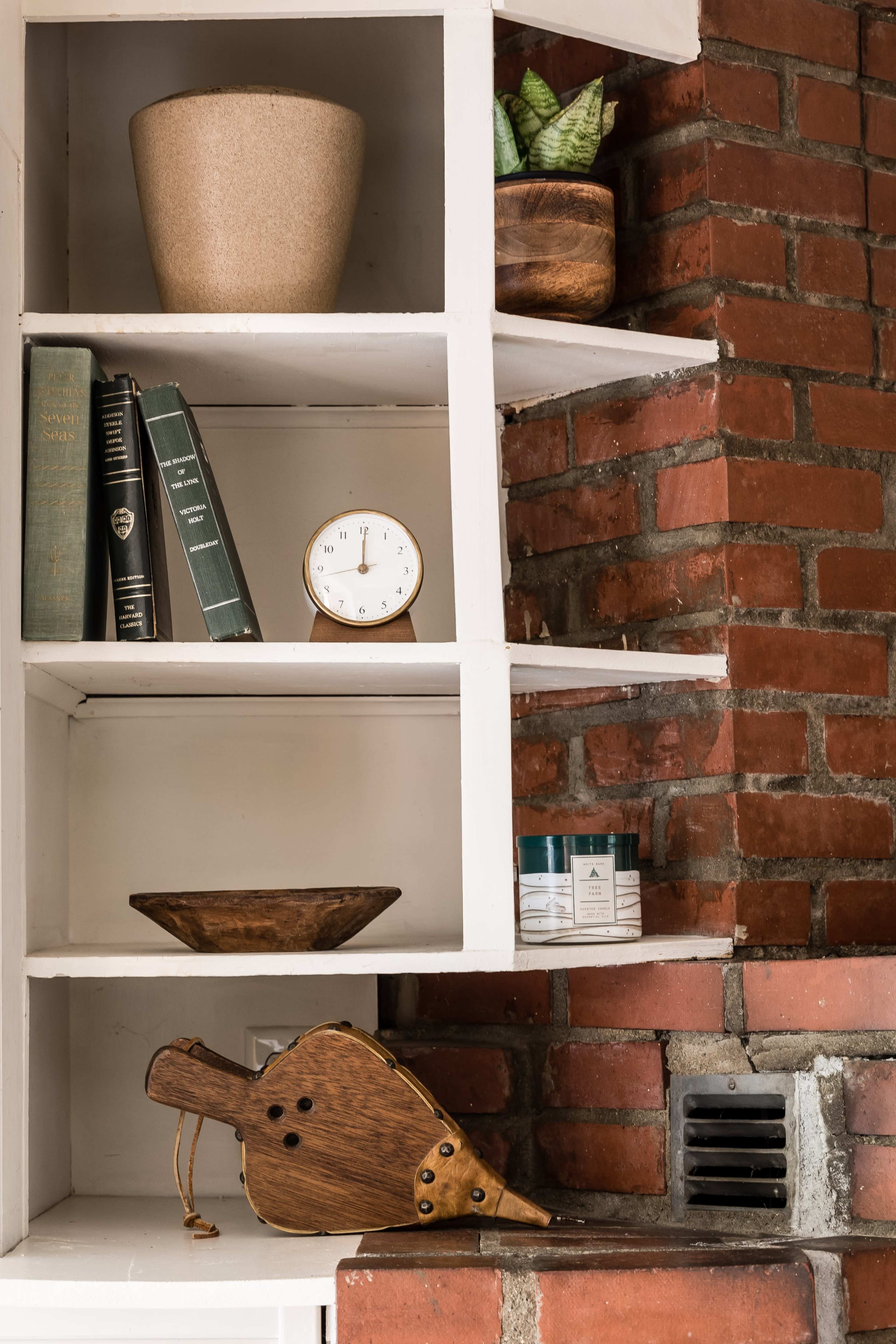 The image shows a shelf with books, a clock, wooden bowls, a plant, and a wooden bellows against a brick wall.