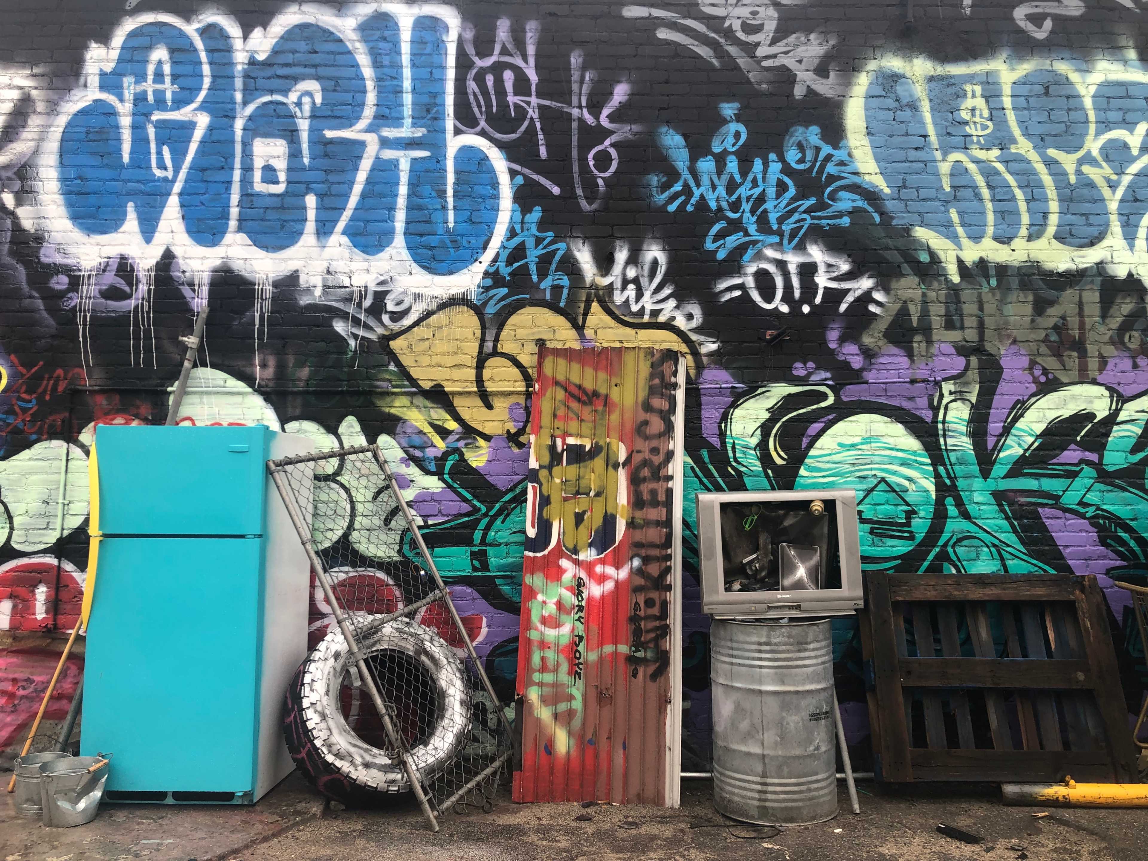 The image shows a colorful graffiti-covered brick wall with various discarded items, including a turquoise refrigerator, a metal door, a tire, and a barrel.