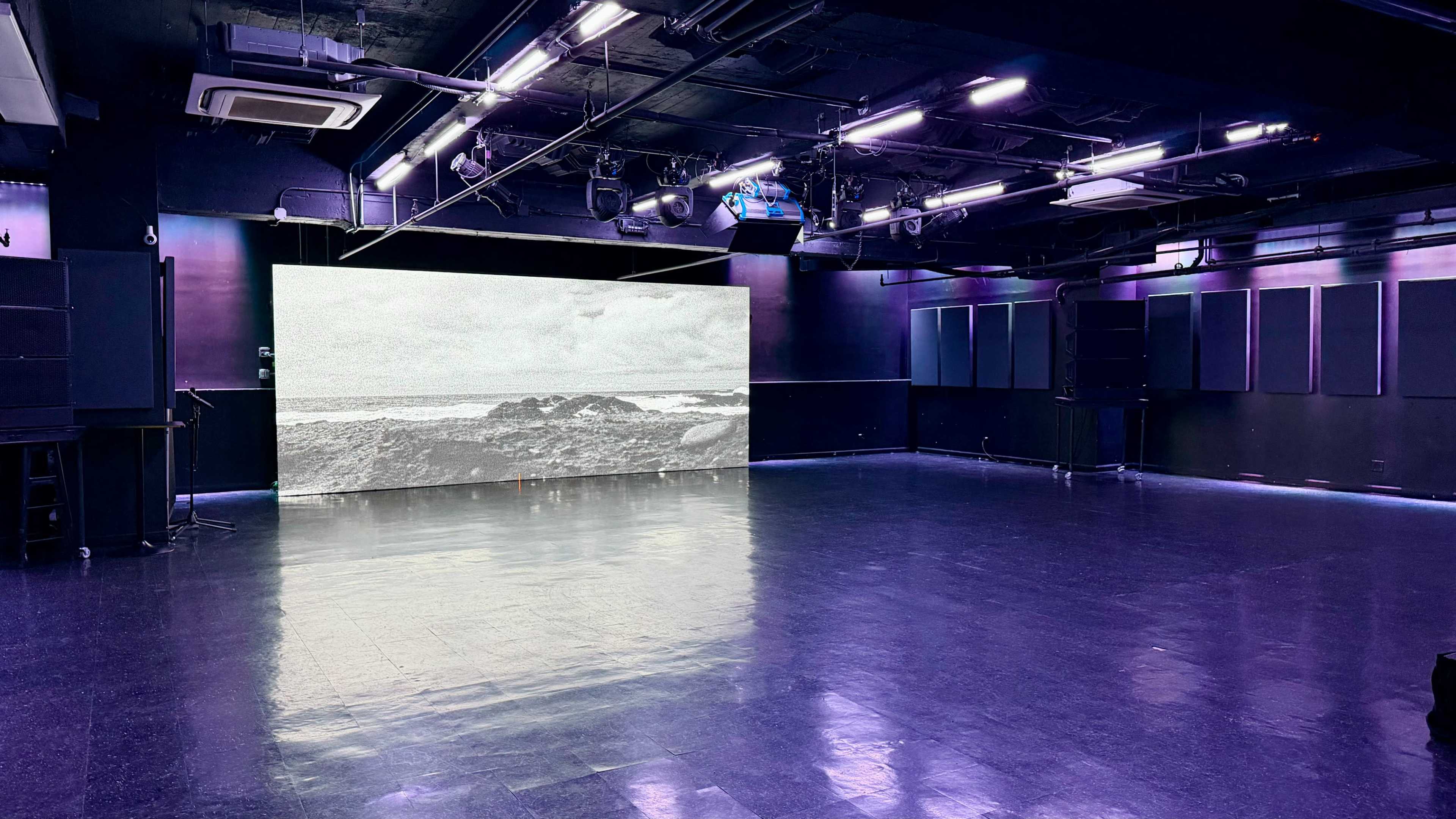 The image shows a dark room with a large screen displaying a black and white coastal scene, surrounded by black walls and illuminated by purple lights.