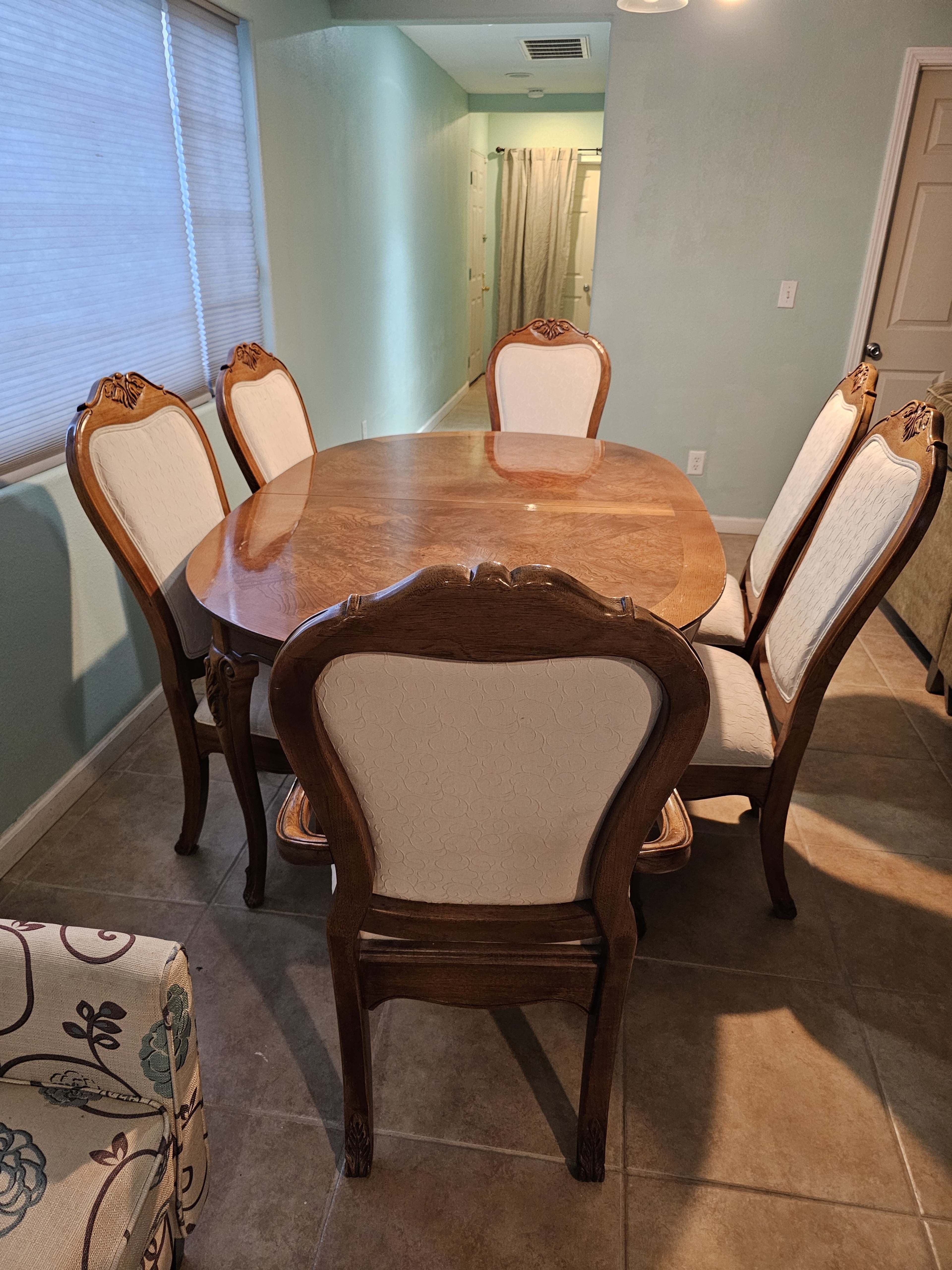 A wooden dining table surrounded by six chairs with upholstered backs is positioned in a room with light-colored walls and a visible shower curtain in the background.