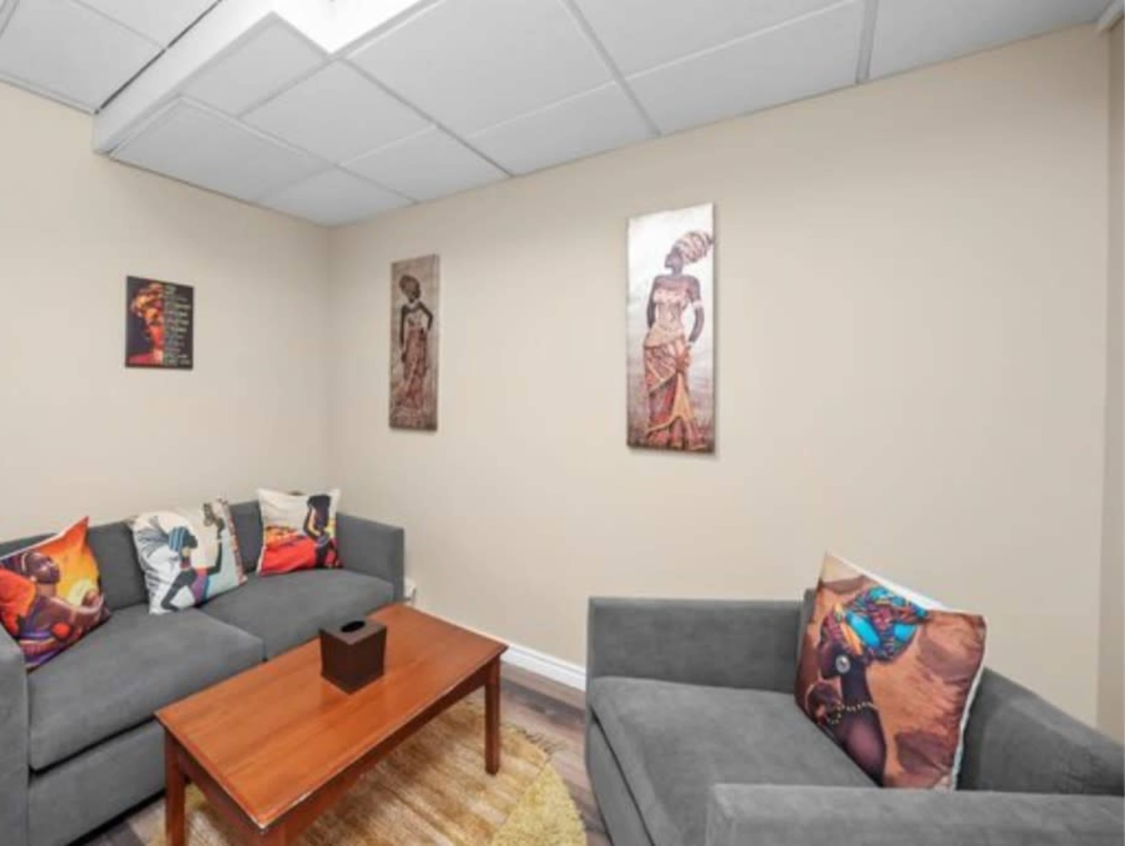 The image shows a small room with two grey sofas, a wooden coffee table in the center, and artwork featuring figures on the walls.