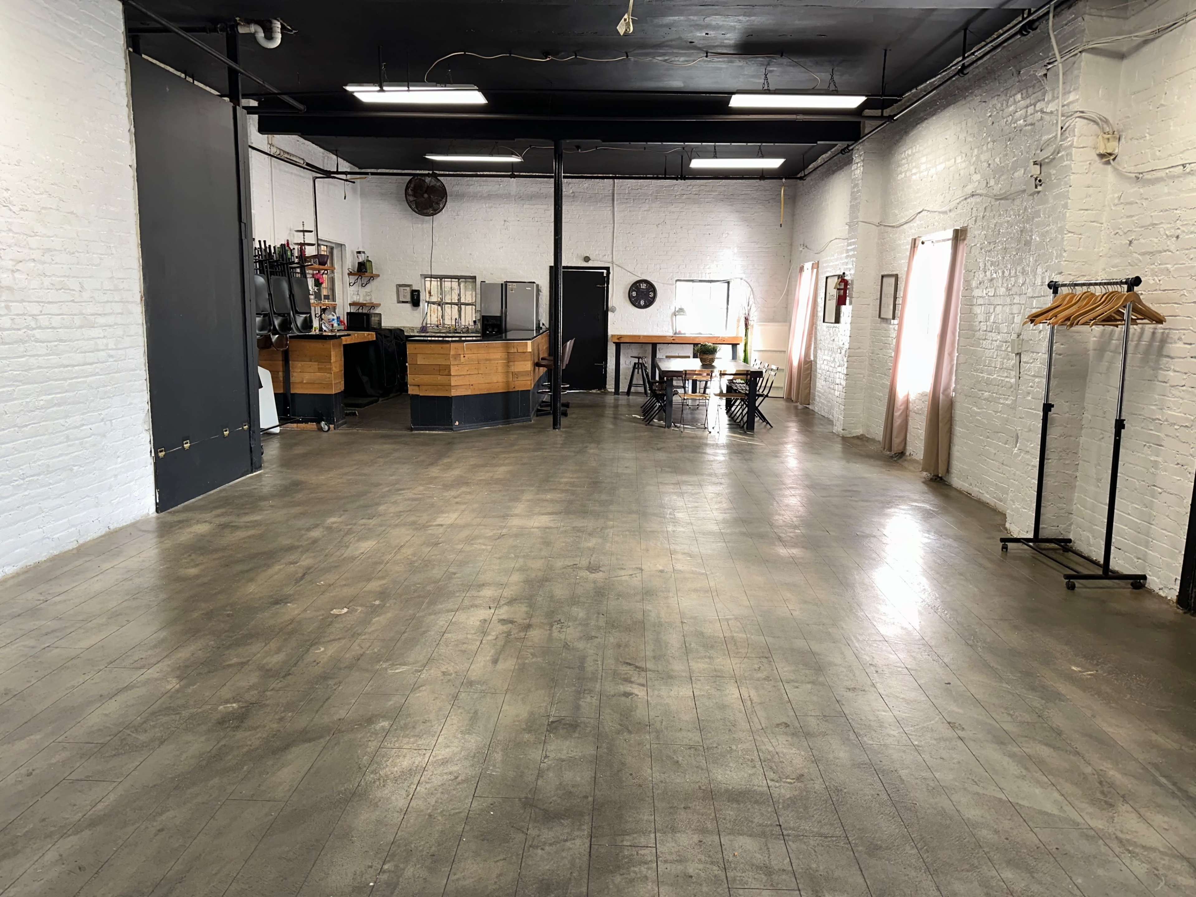The image shows an empty indoor space with wooden flooring, exposed brick walls, and a small bar area in the background.