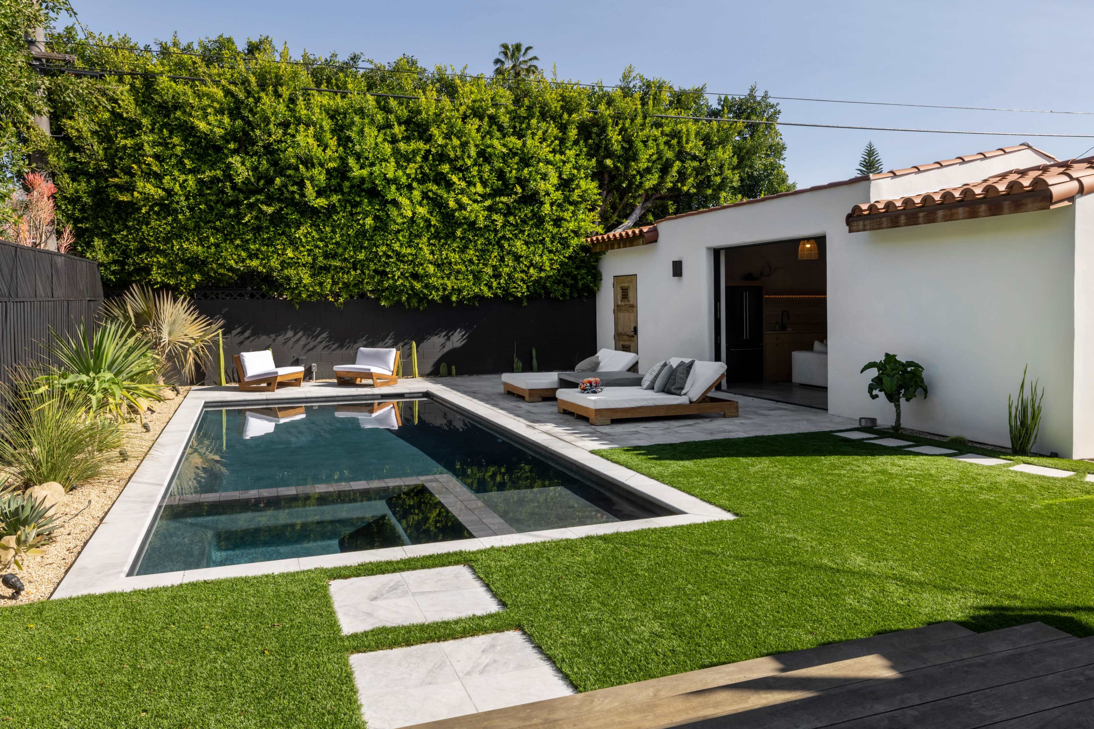 A modern outdoor space featuring a rectangular swimming pool surrounded by lounge chairs, artificial turf, and a wall of greenery.