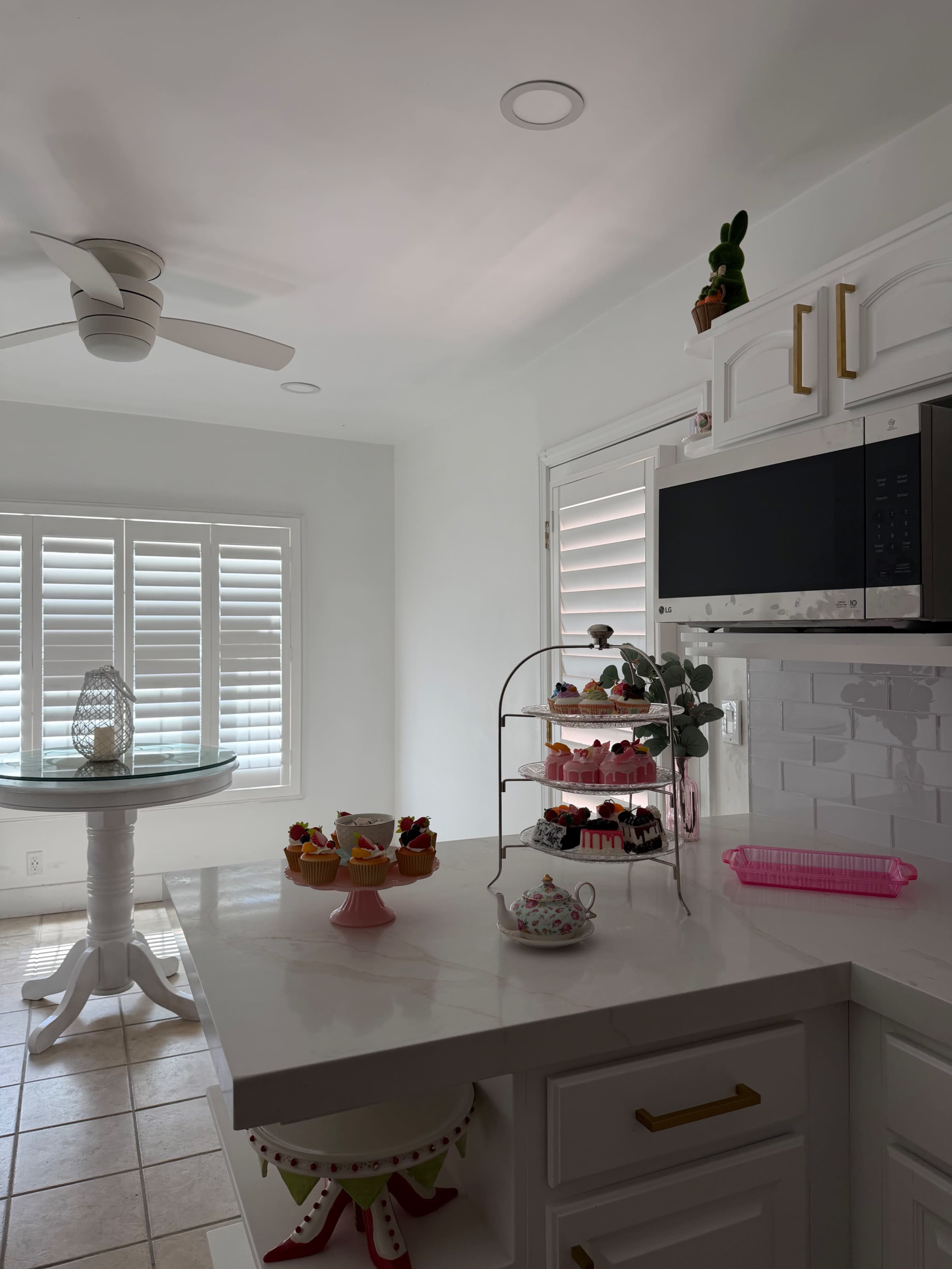 A bright kitchen features a round table with a glass top, a tiered dessert stand filled with sweets, and a microwave above a white countertop with a bowl of fruit.