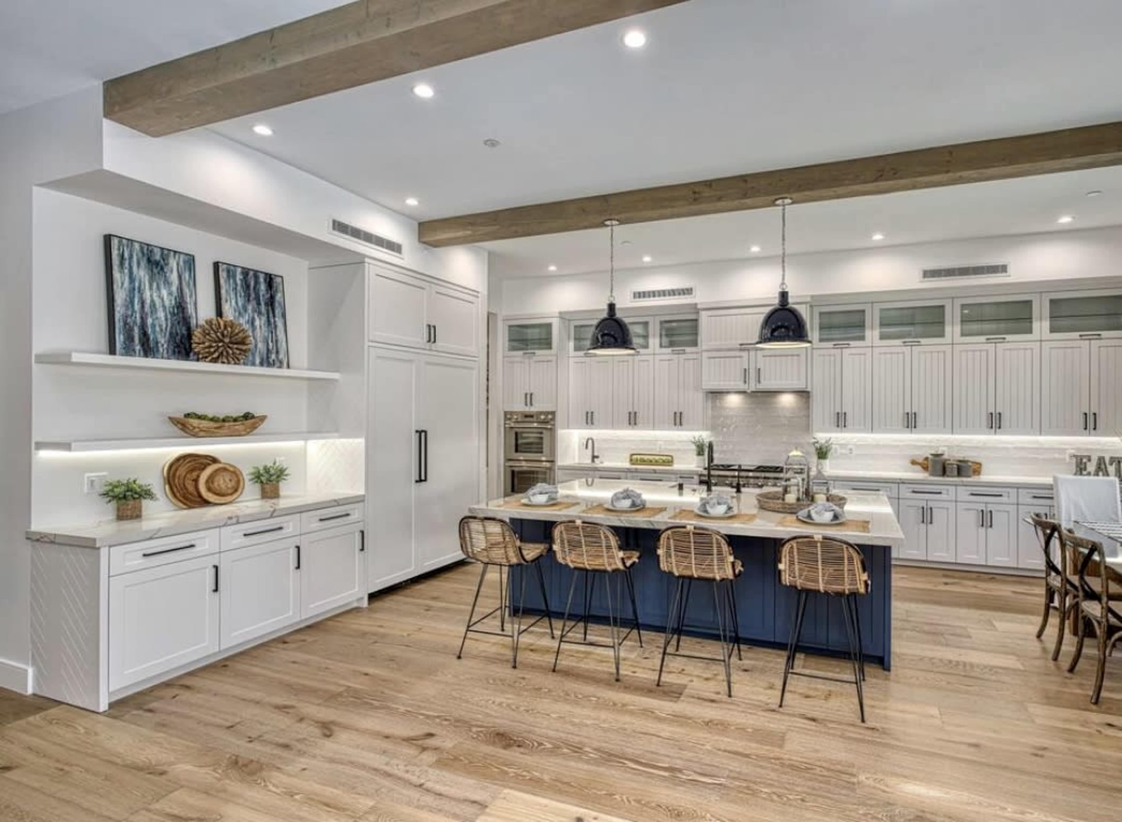 The image shows a modern kitchen with white cabinetry, a blue island, and wooden beams overhead, featuring barstools and contemporary lighting.