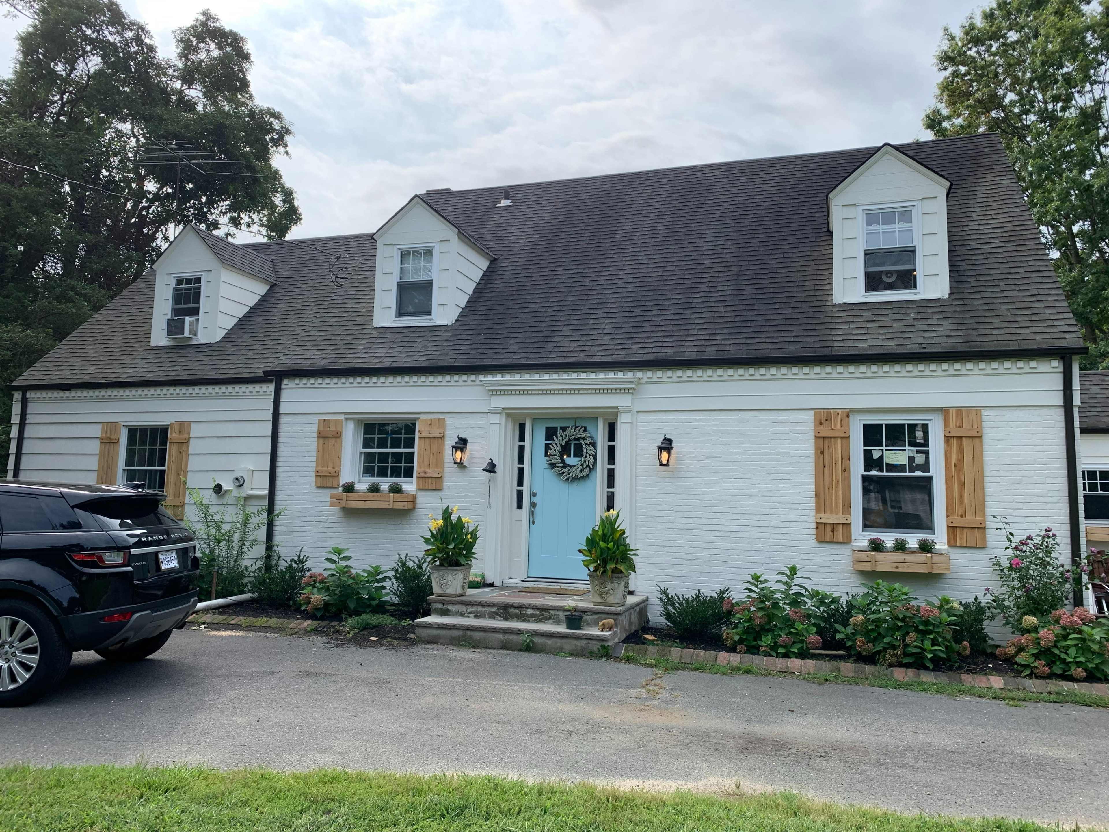 A two-story, white house with a blue door and wooden shutters, surrounded by a landscaped garden, is parked in front of a black car.