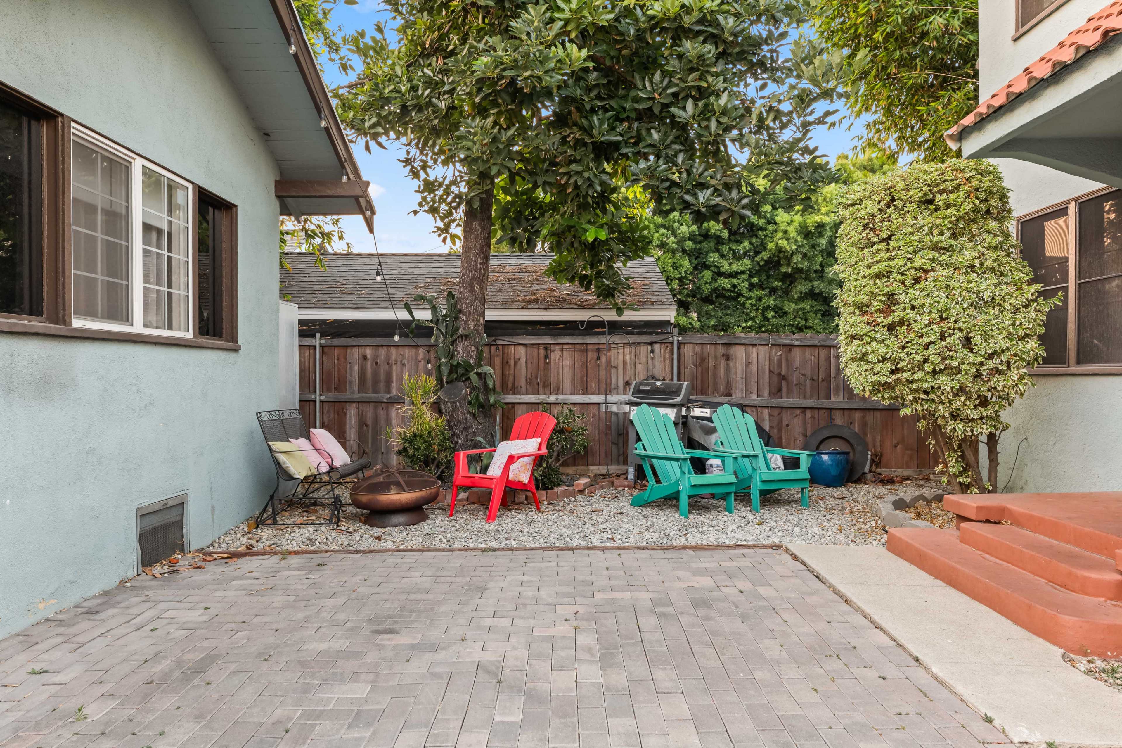 The image shows a small outdoor patio area featuring colorful chairs, a grill, and landscaping with a tree and shrubs.