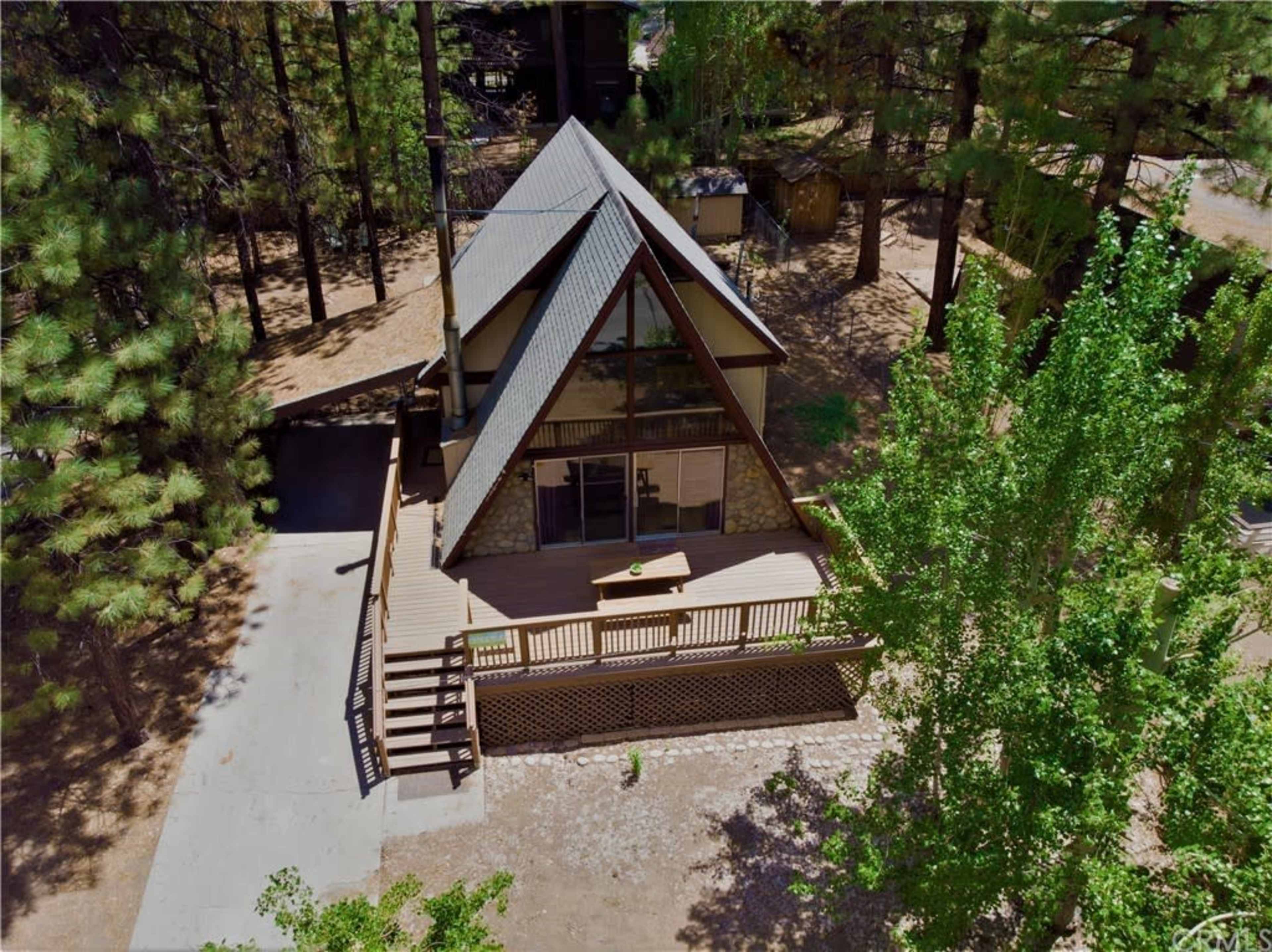 Aerial view of a two-story A-frame cabin surrounded by trees, featuring a large deck and a gravel driveway.