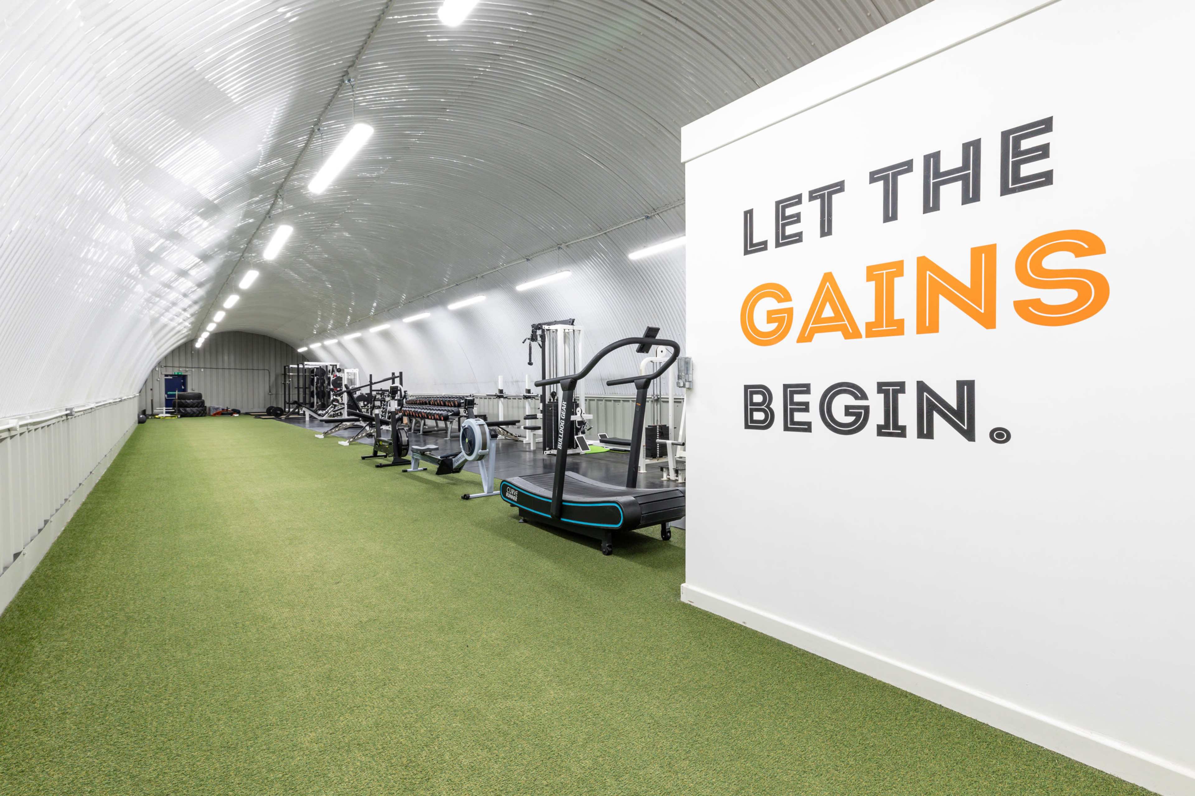 The image shows a long, narrow gym space with artificial turf flooring and various exercise equipment lined along the walls.