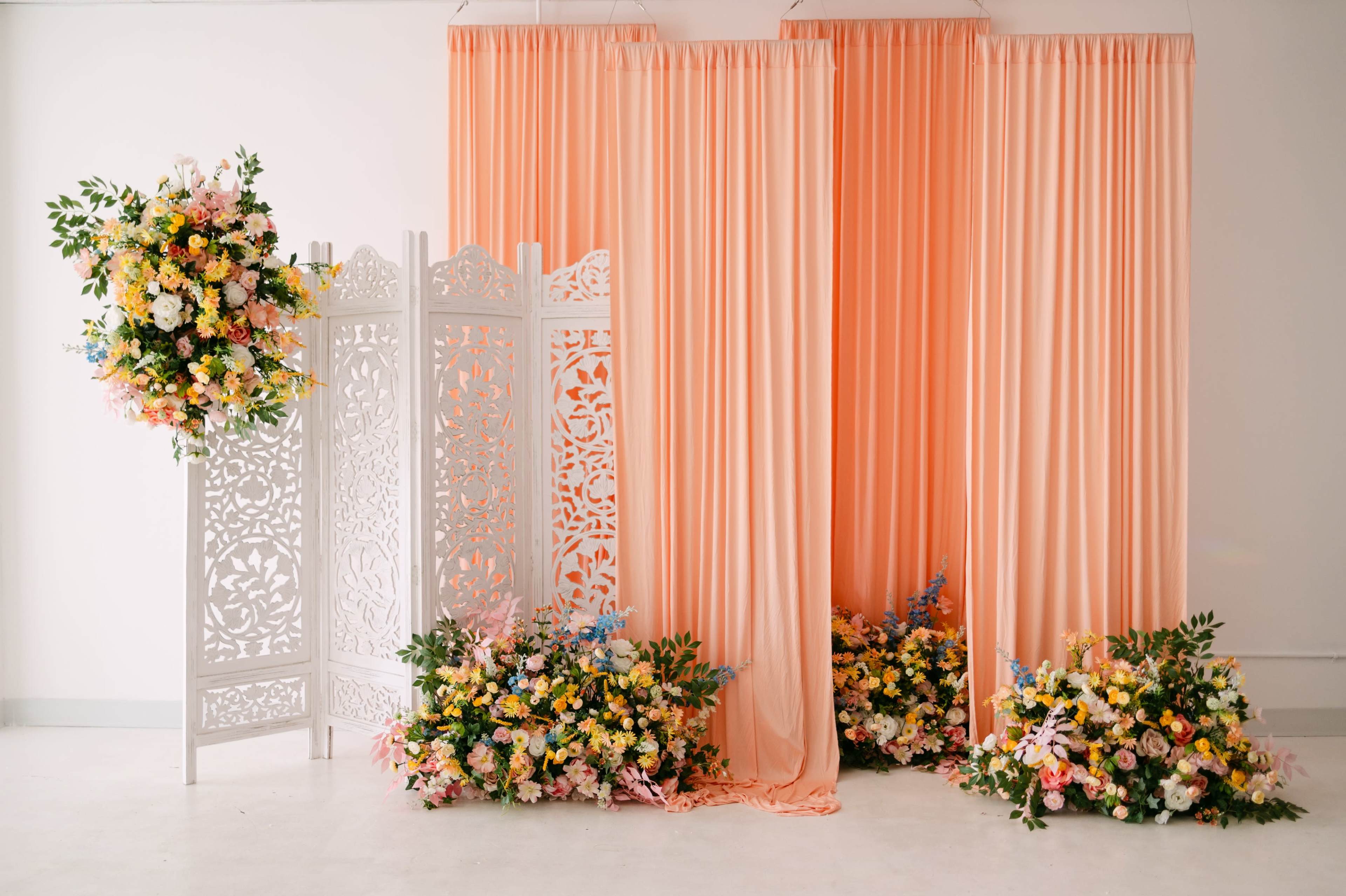 The scene features a backdrop of soft peach curtains, a decorative white screen, and an arrangement of colorful floral decorations on the floor.