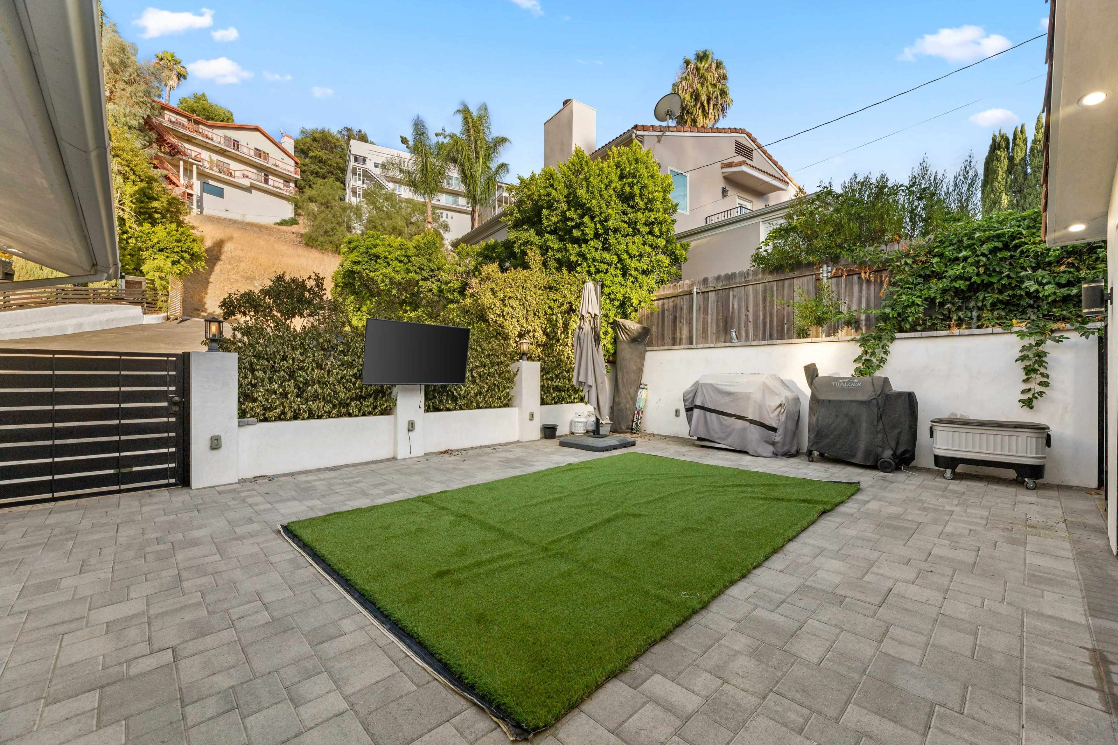 The image shows a paved outdoor area with artificial grass, a television mounted on the wall, a barbecue grill under a cover, and surrounding greenery.