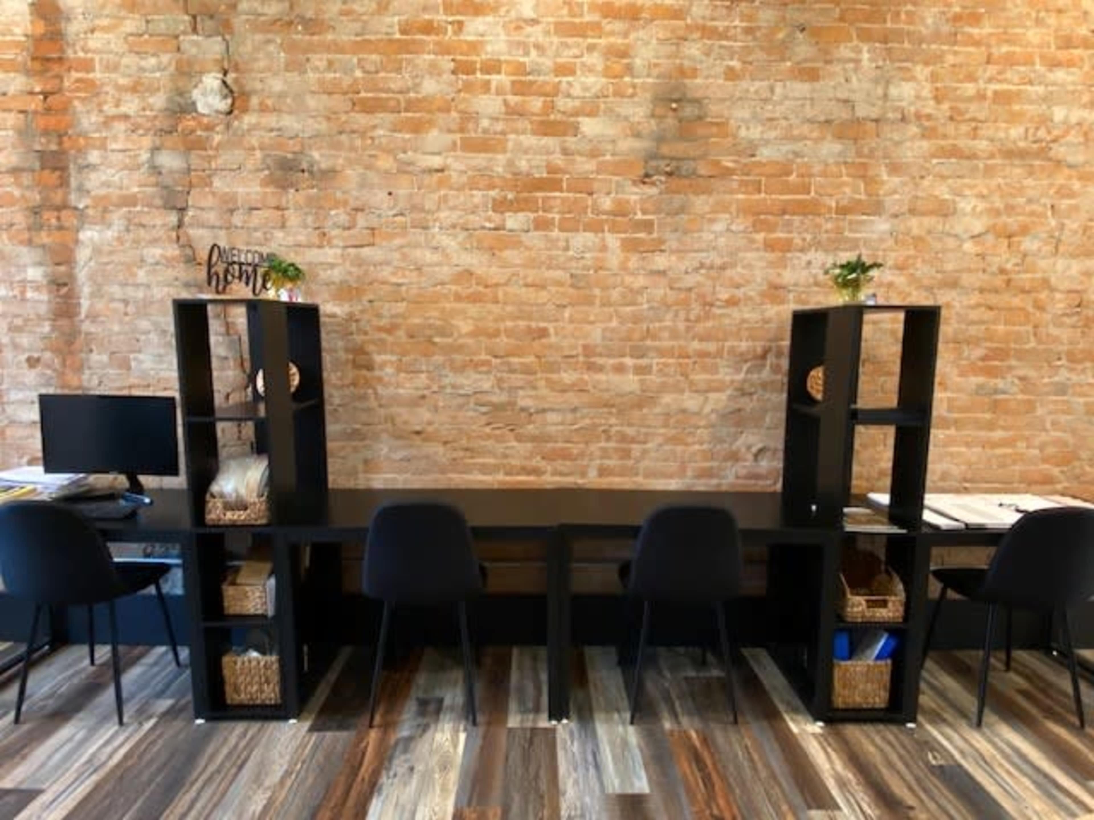 A long, black wooden desk with black chairs is set against a rustic brick wall, flanked by two black shelving units containing baskets and decorative items.