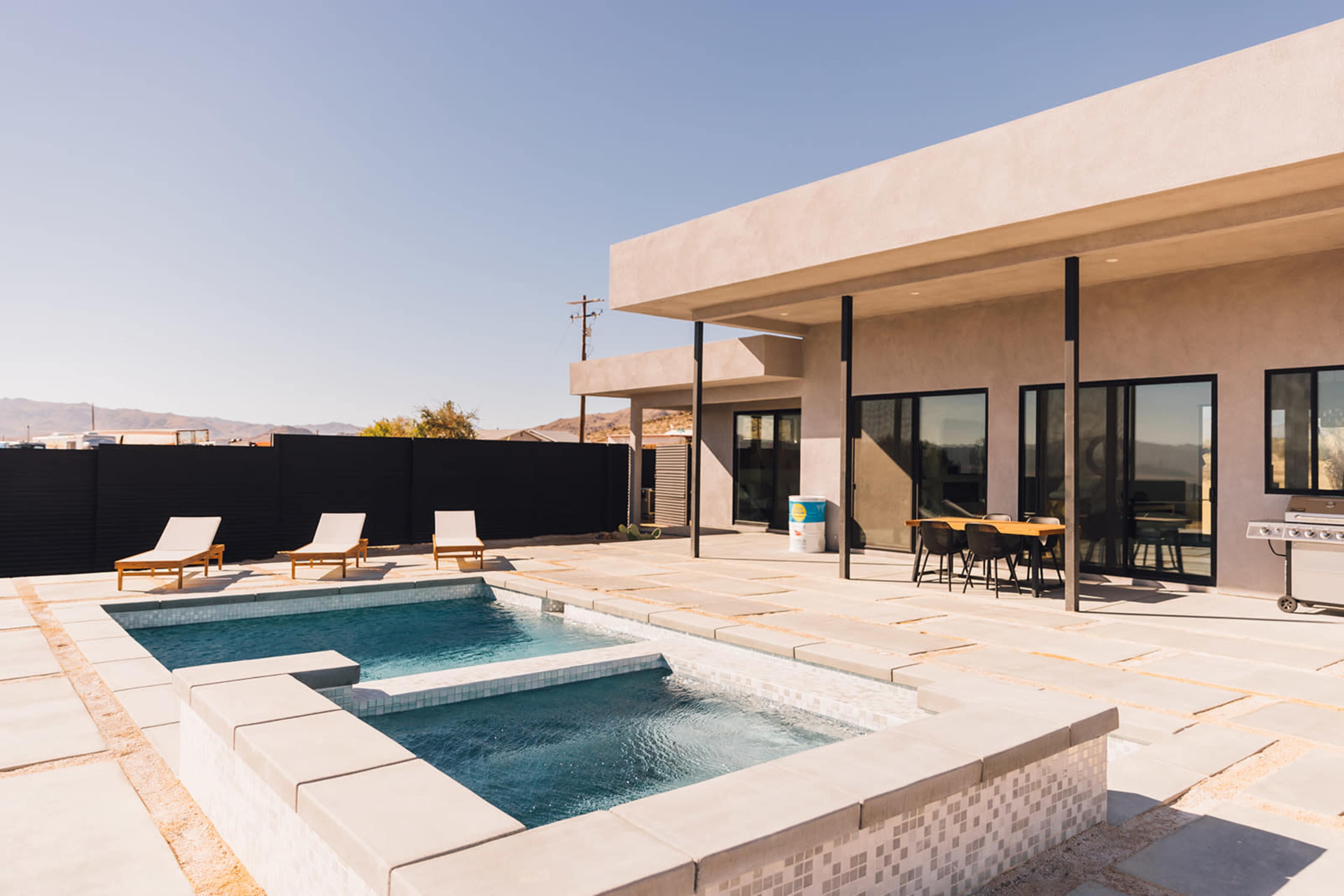 A contemporary outdoor space featuring a pool, jacuzzi, lounge chairs, and a shaded dining area.