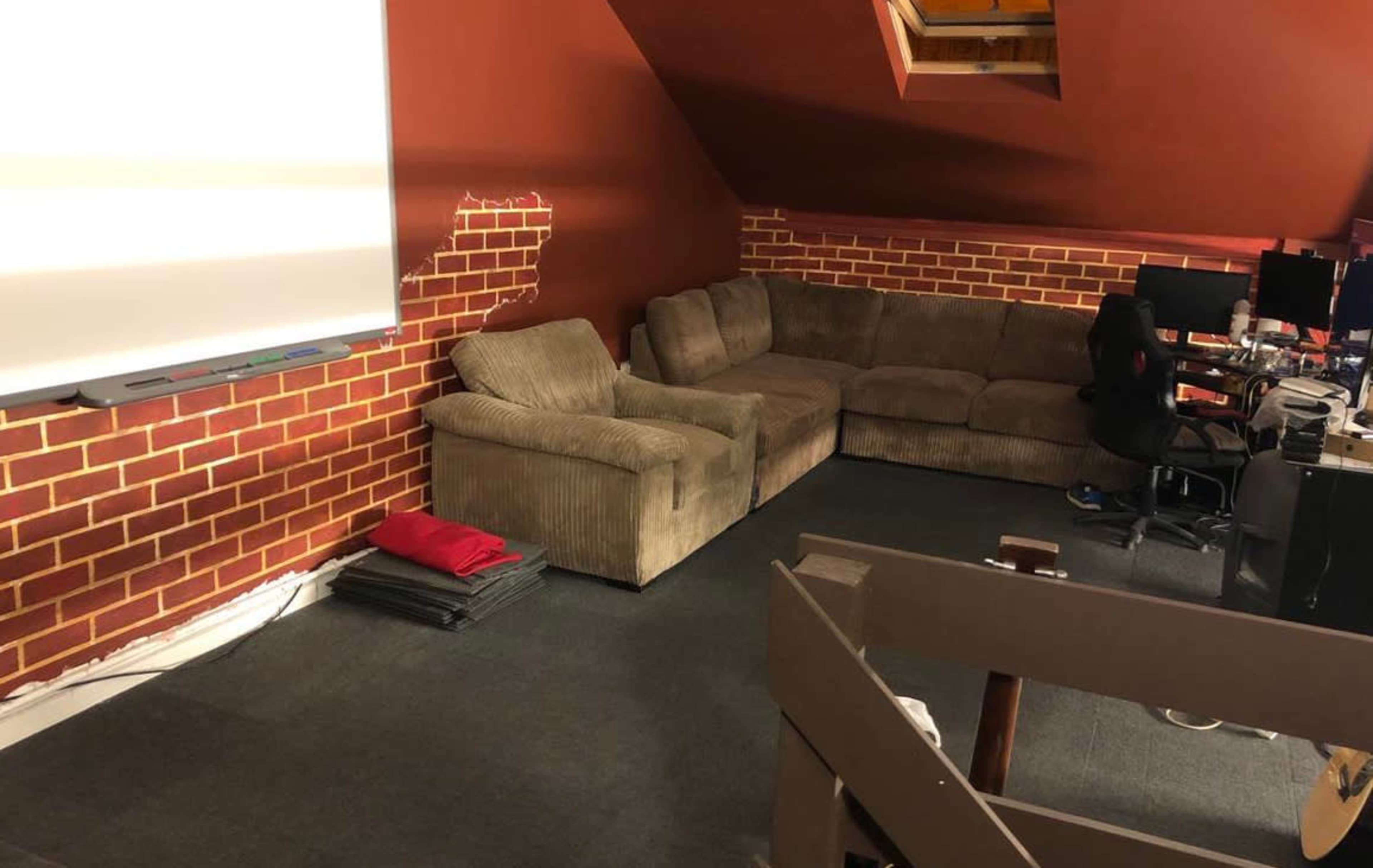 A spacious attic room features a brown sectional couch, a flat-screen projector screen, and workstations with computers.