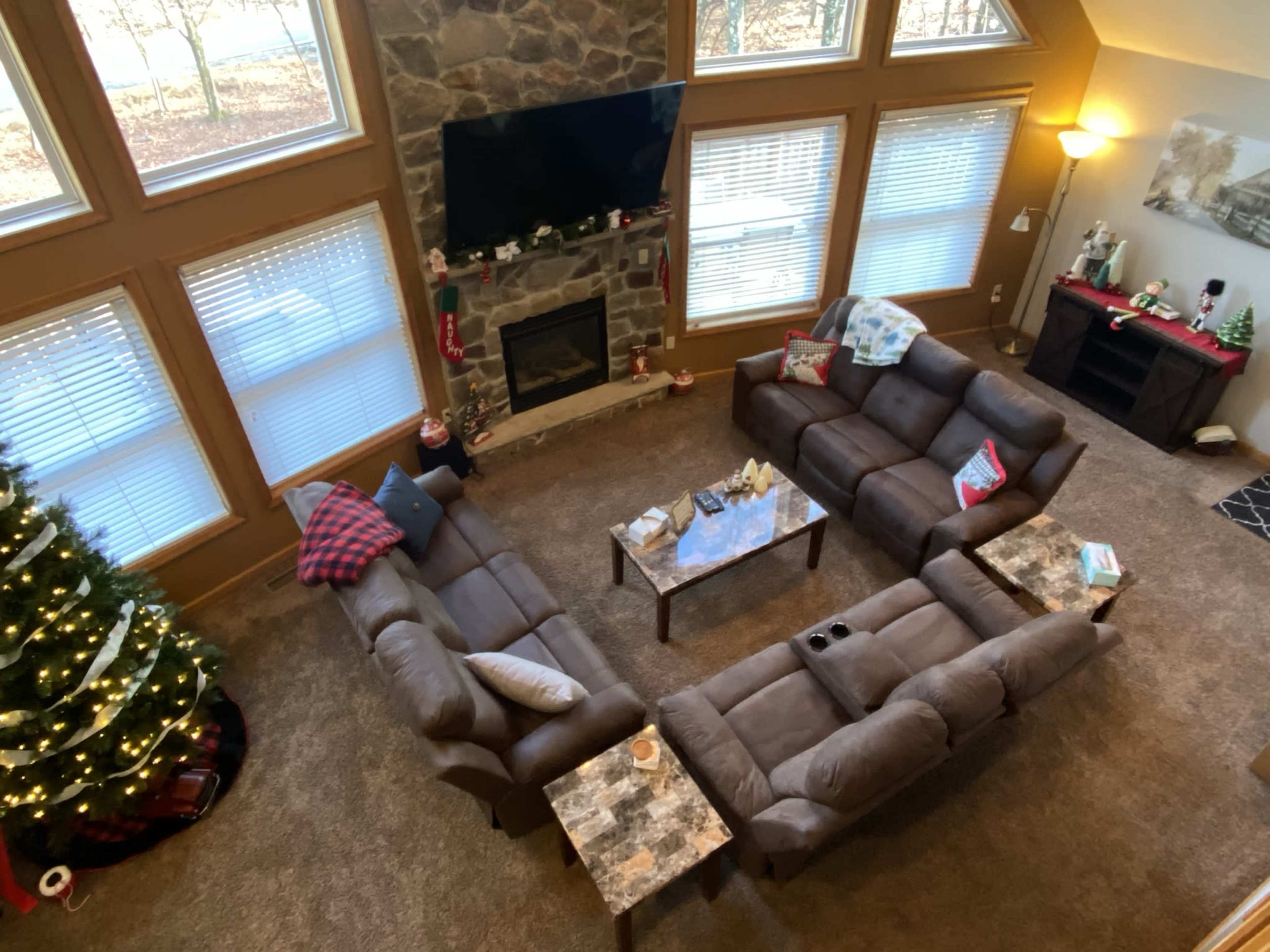 A spacious living room with two brown couches arranged in a U-shape around a coffee table, a stone fireplace, and a Christmas tree in the corner.