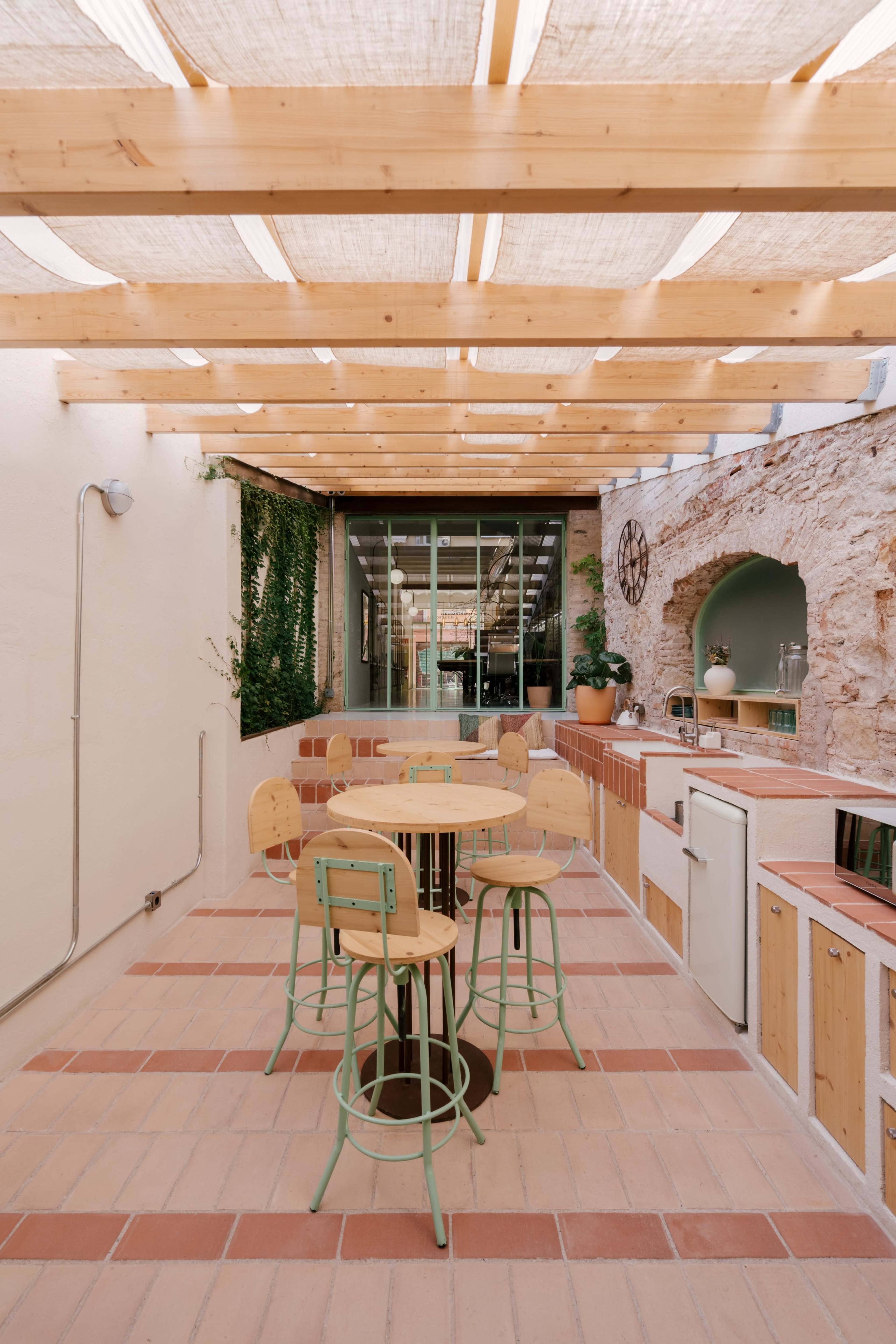 The image shows a modern outdoor patio with wooden beams overhead, featuring a round table and green metal stools beside a kitchen area.
