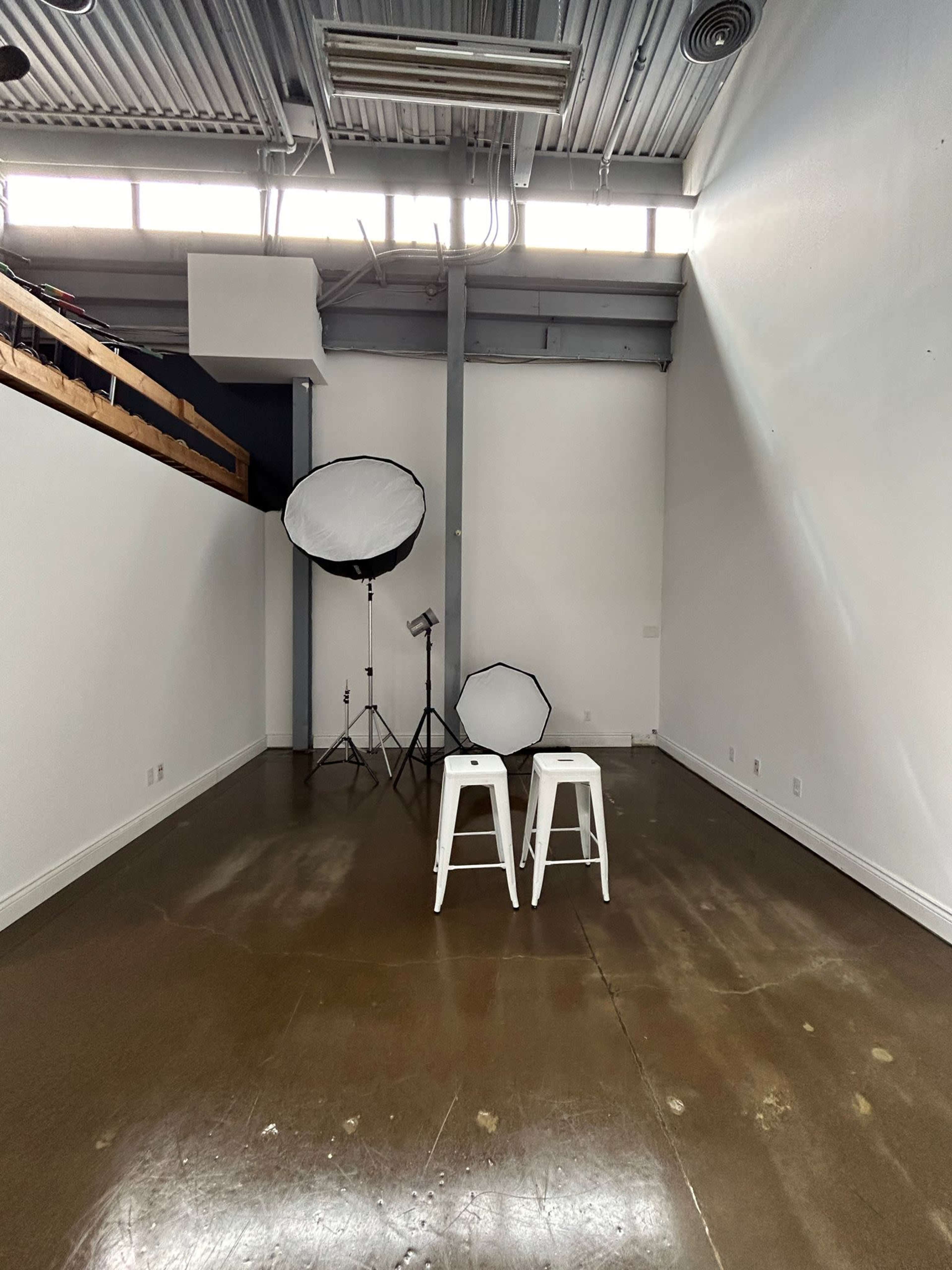 The image shows a spacious, minimalist studio with two white stools and photography equipment positioned against plain walls.