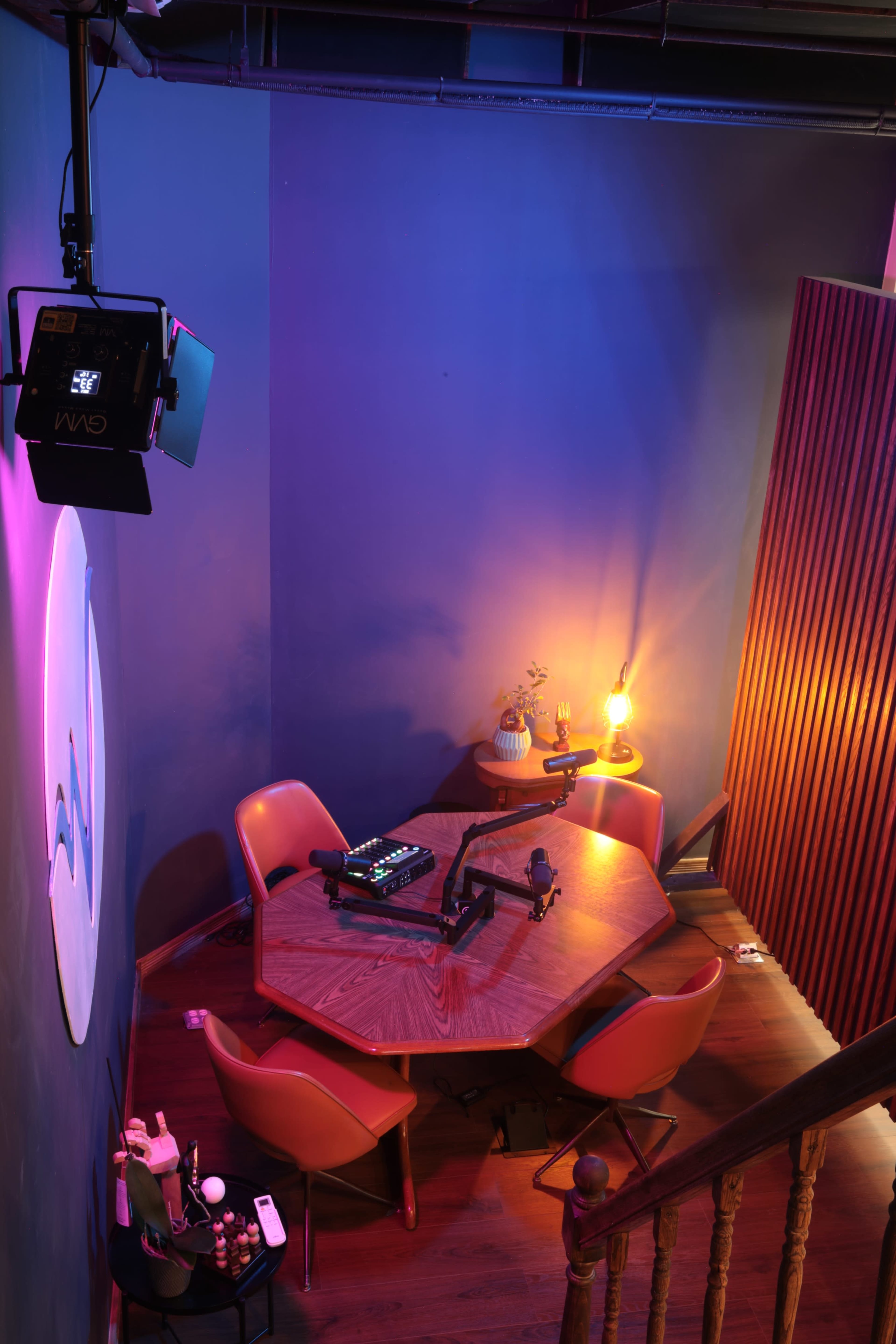A dimly lit studio features a hexagonal wooden table surrounded by four orange chairs, with a spotlight and decorative items on display.