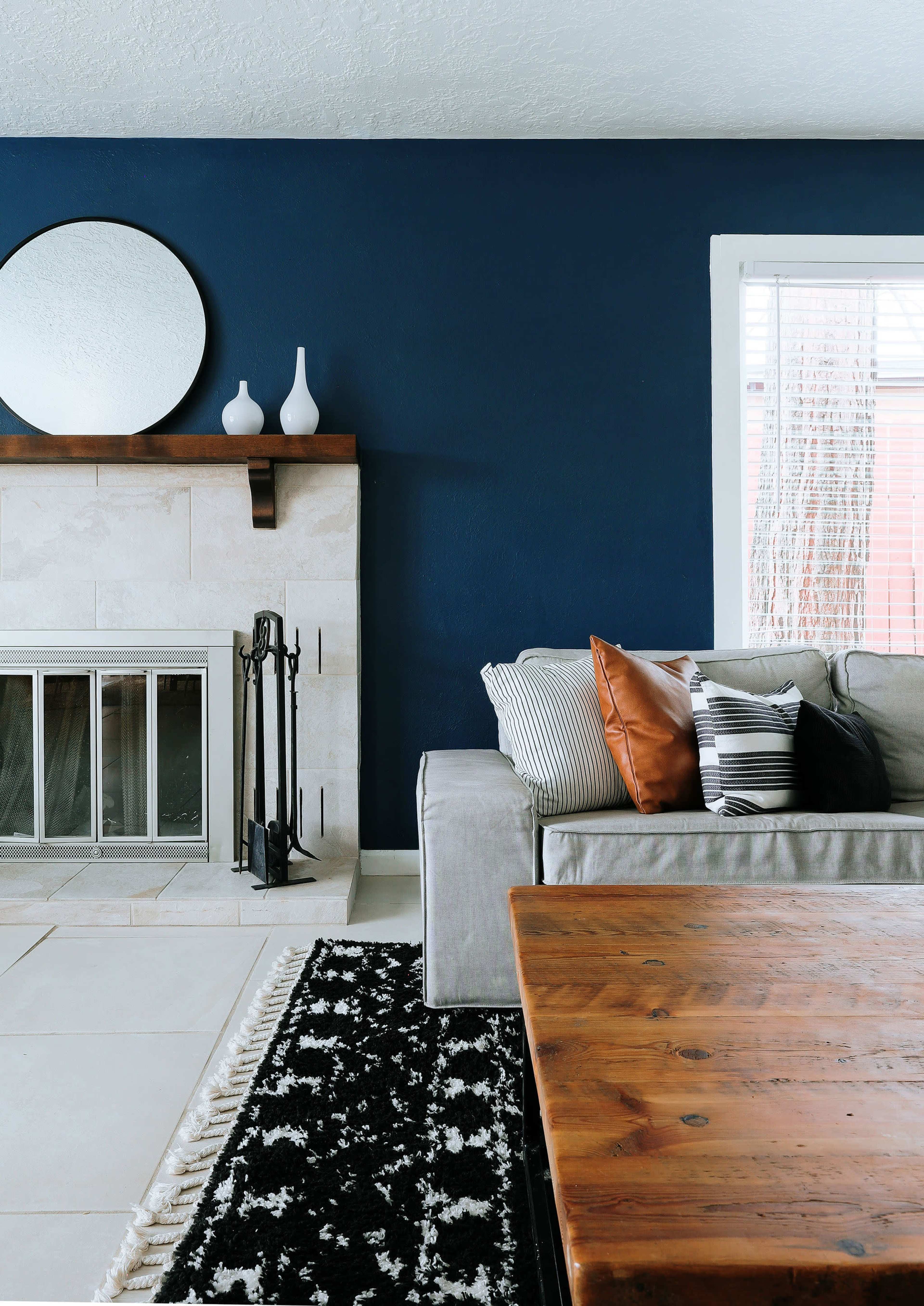 A living room with a dark blue wall, a stone fireplace, a gray sofa adorned with decorative pillows, and a wooden coffee table on a black and white patterned rug.