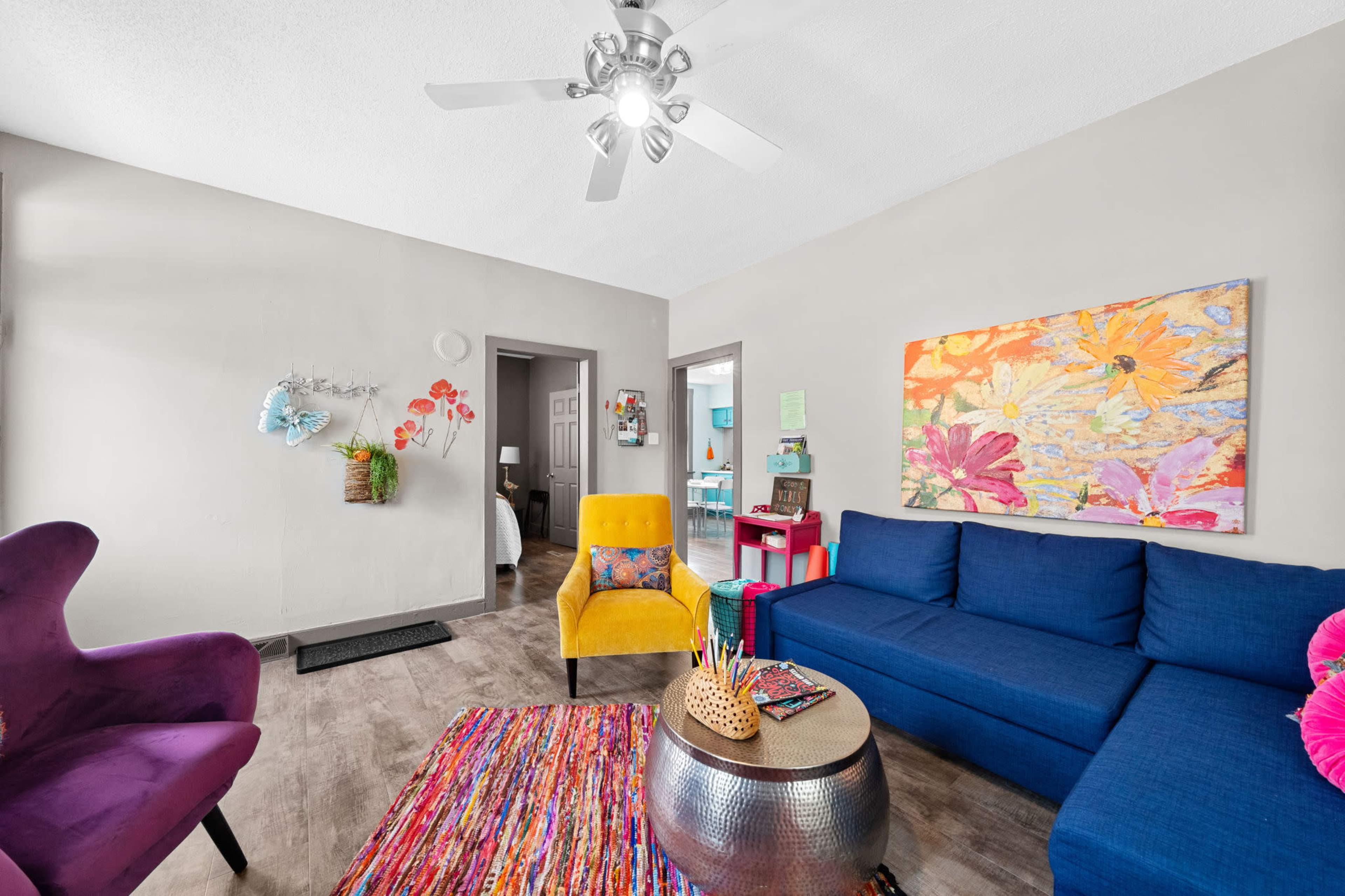 The living room features a blue couch, a yellow chair, a colorful area rug, and a wall art display with floral designs.
