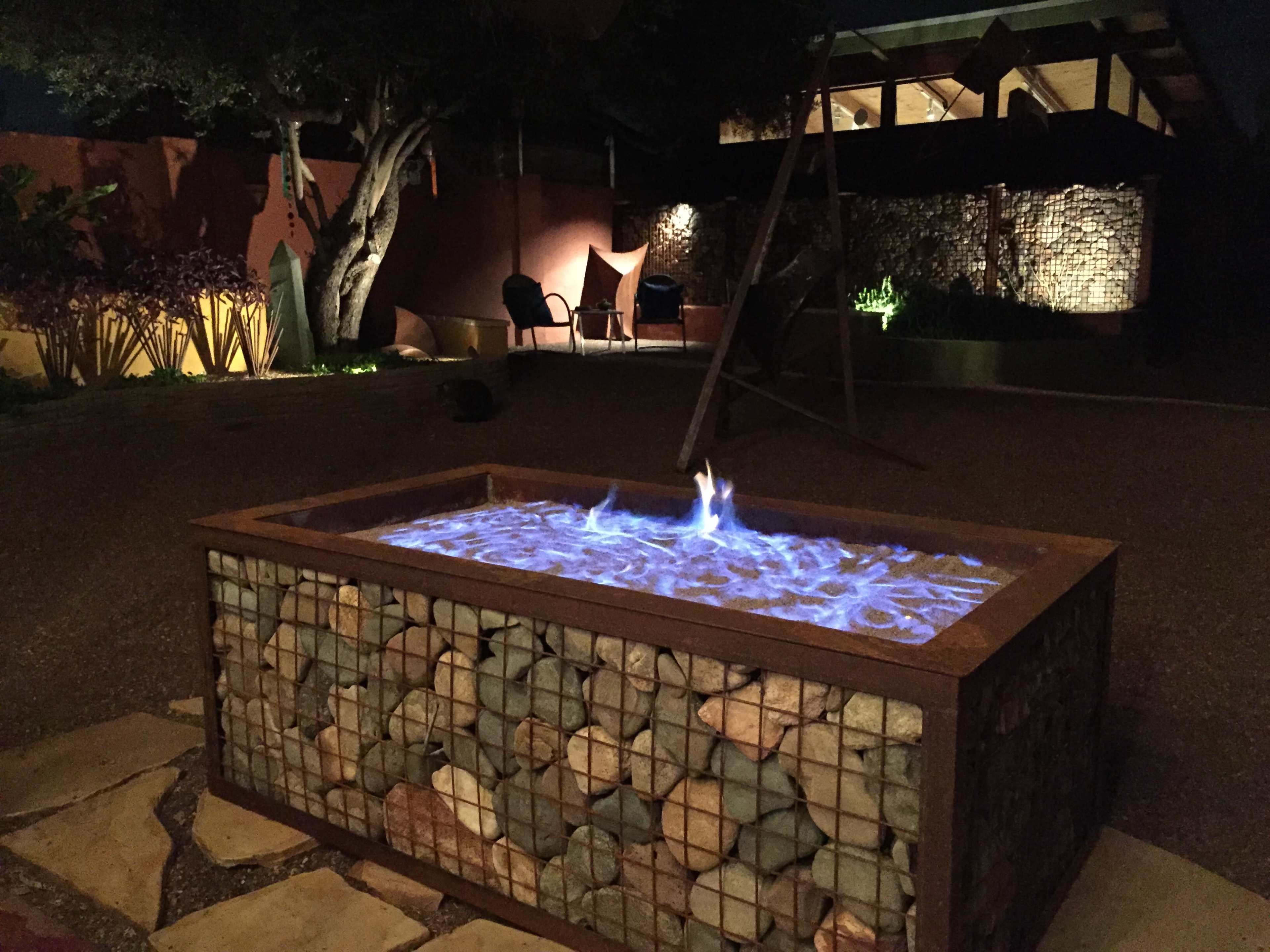 A stone and metal fire pit with blue flames sits in a dimly lit outdoor area surrounded by landscaping and seating.