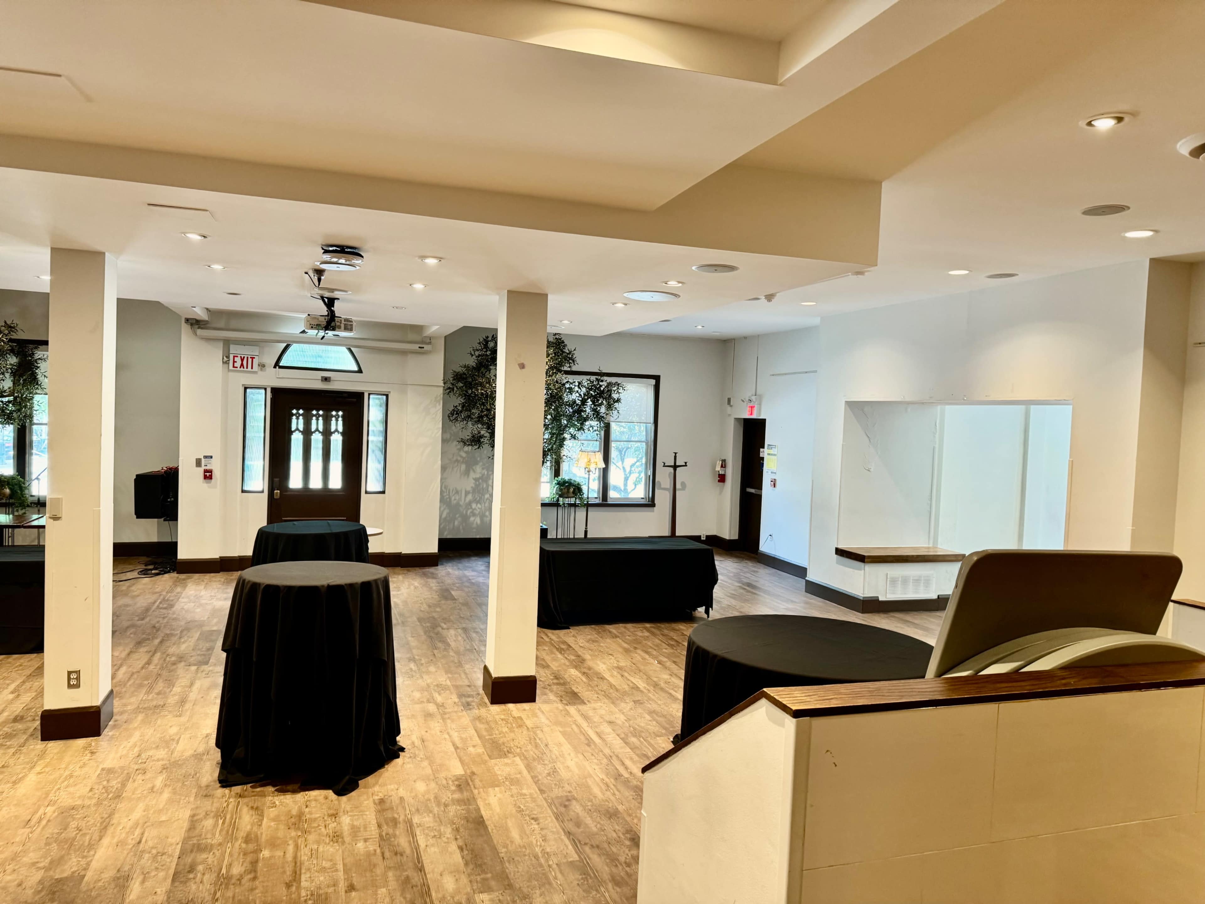The image shows a spacious, well-lit event room featuring several round tables covered in black cloth and large windows with natural light.