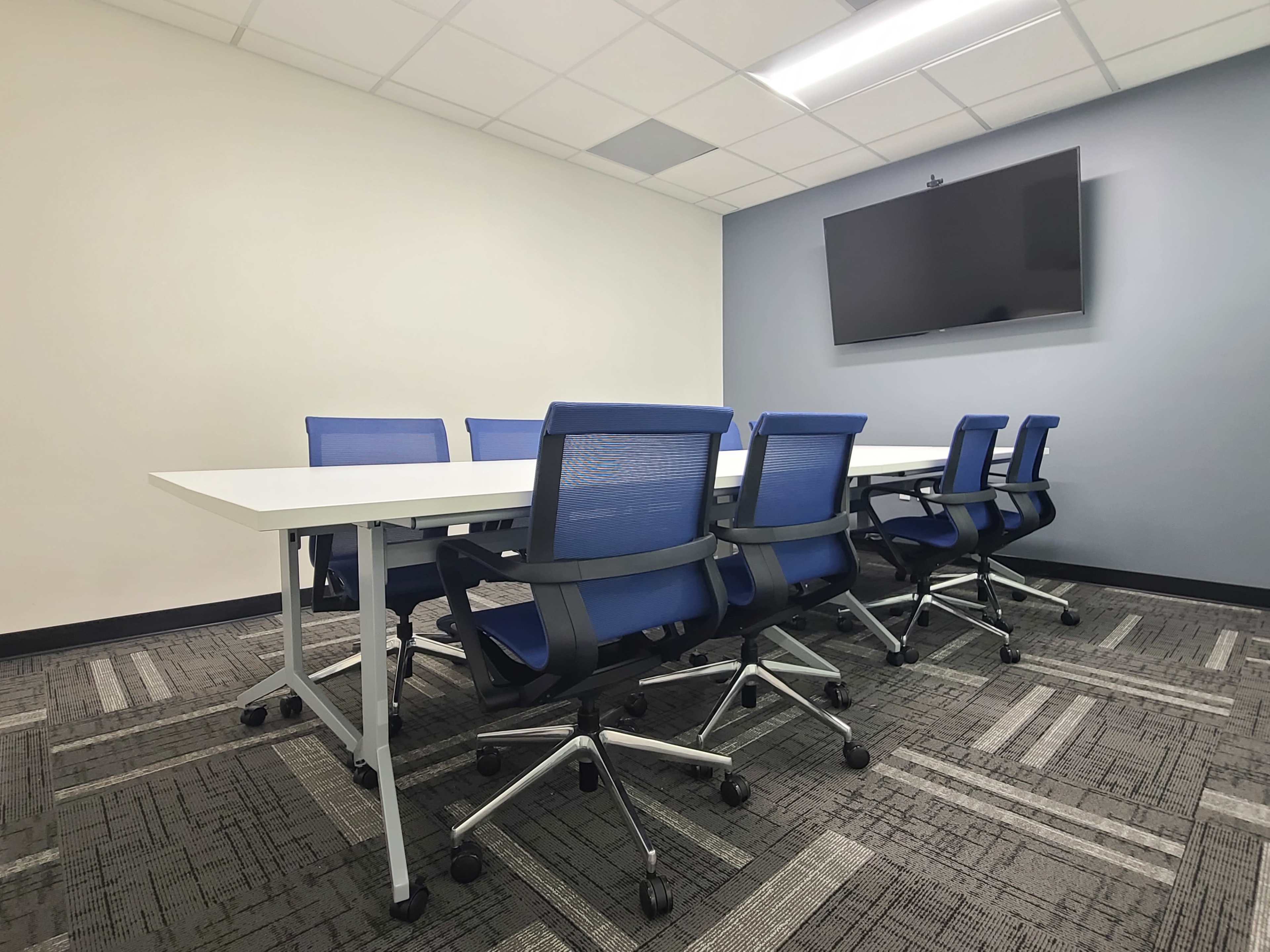 A modern conference room features a long white table surrounded by blue ergonomic chairs and a wall-mounted television.