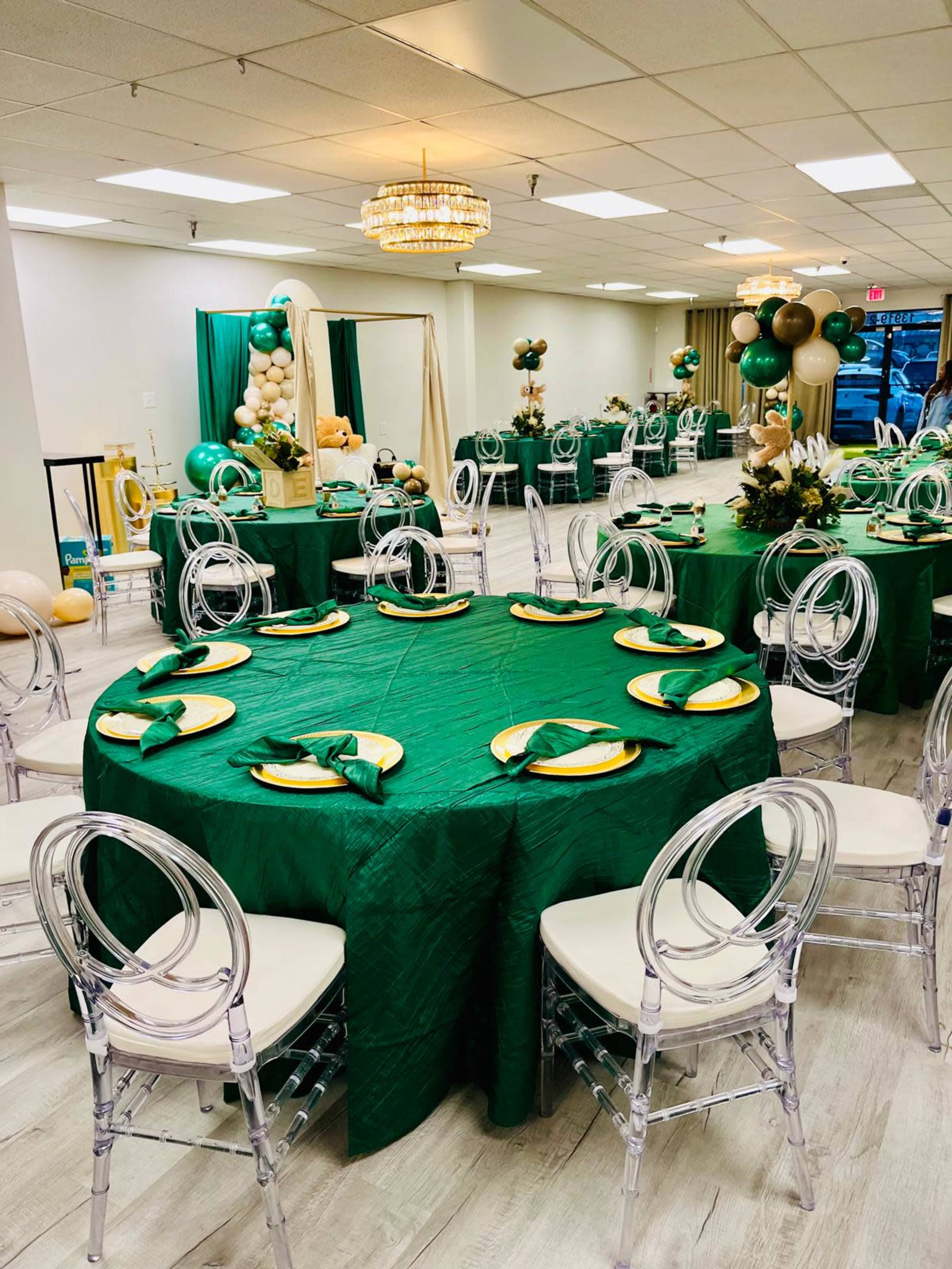 The image shows a banquet hall set up with green tablecloths, clear acrylic chairs, and decorative balloons arranged around the space.