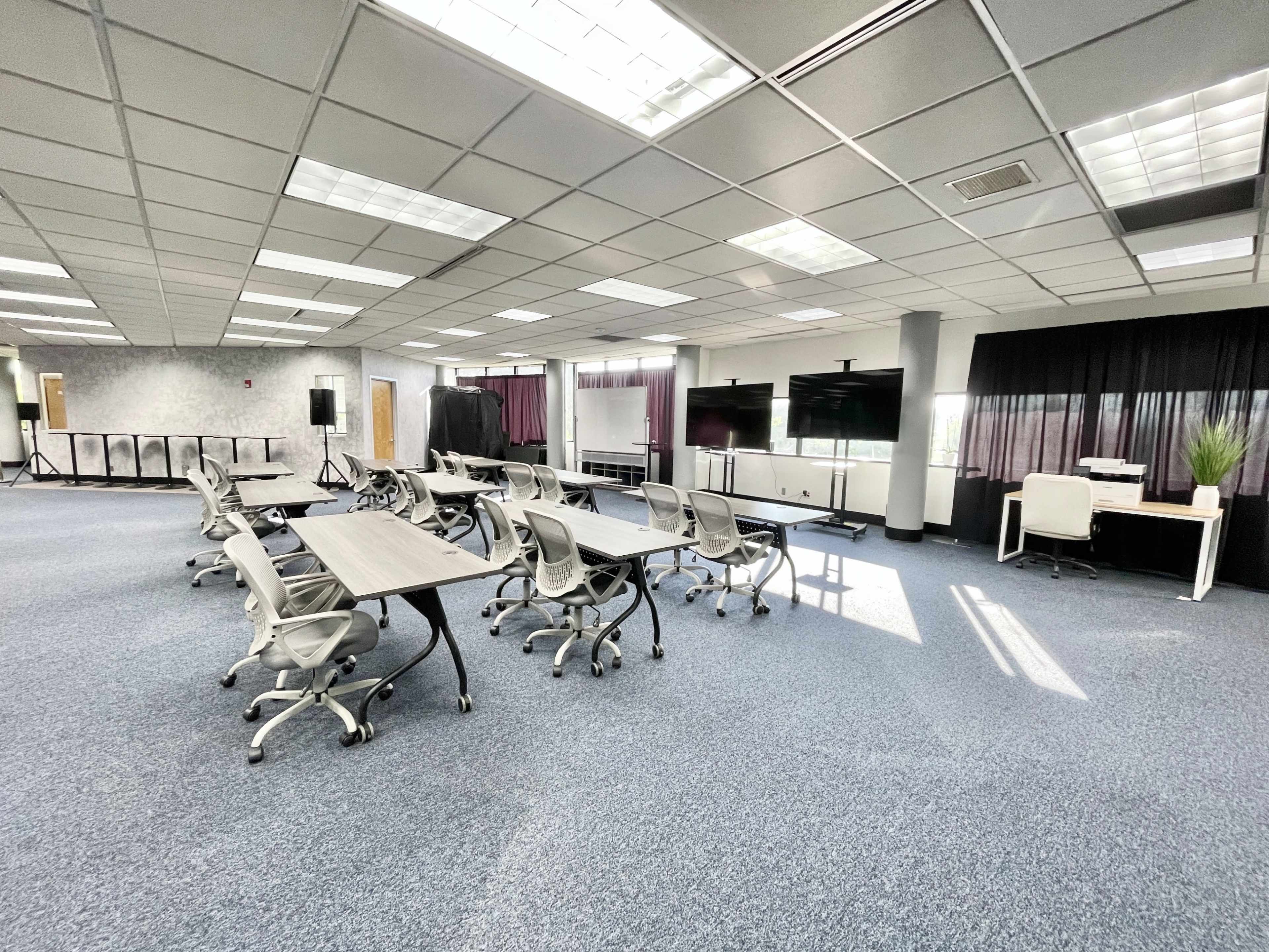 The image shows a spacious office area with several tables and chairs arranged for meetings, illuminated by natural light from overhead windows.