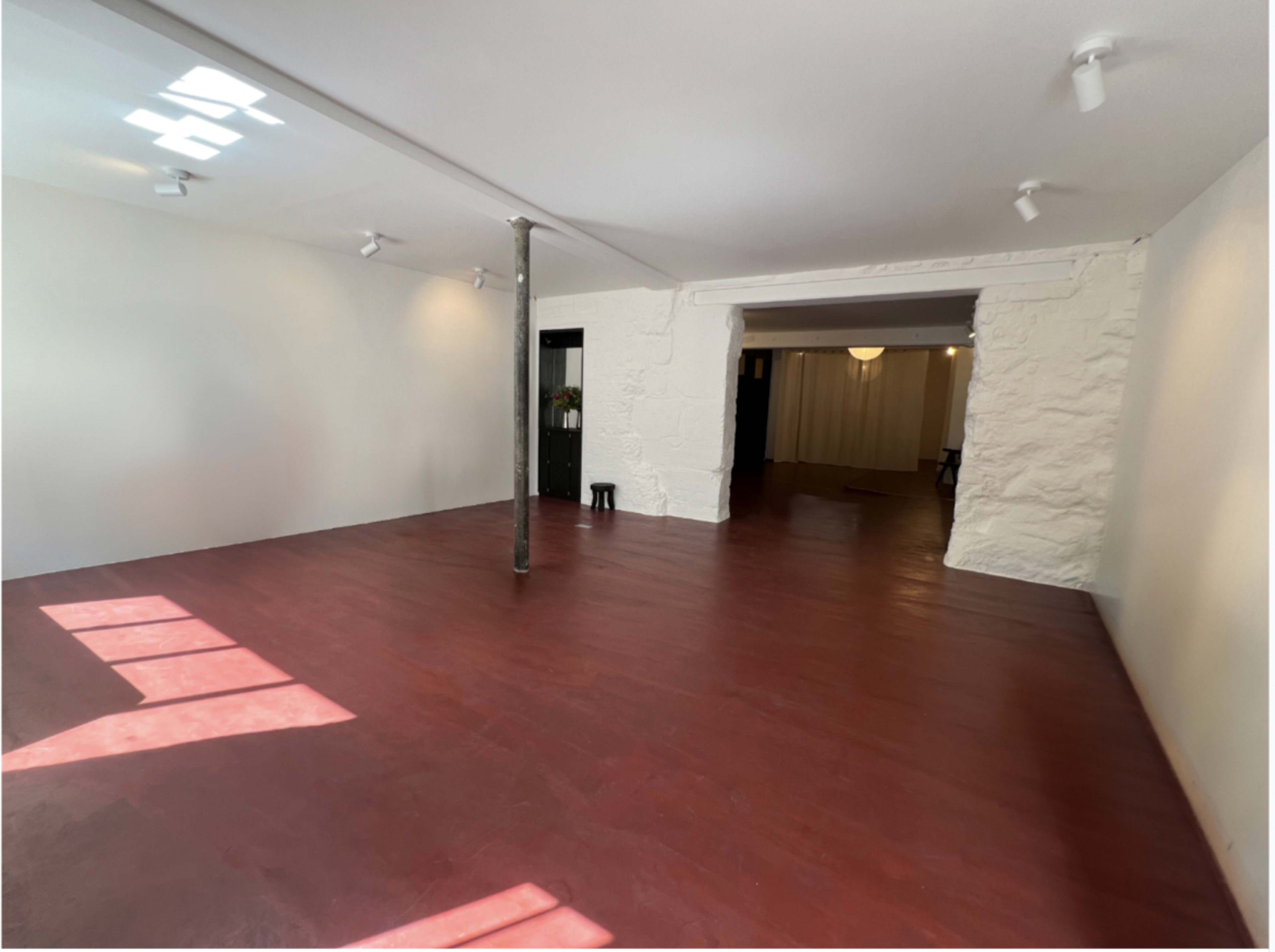 The image shows an empty, well-lit room with a red floor and a column in the center, leading to an adjoining space with white walls and a light-colored curtain.
