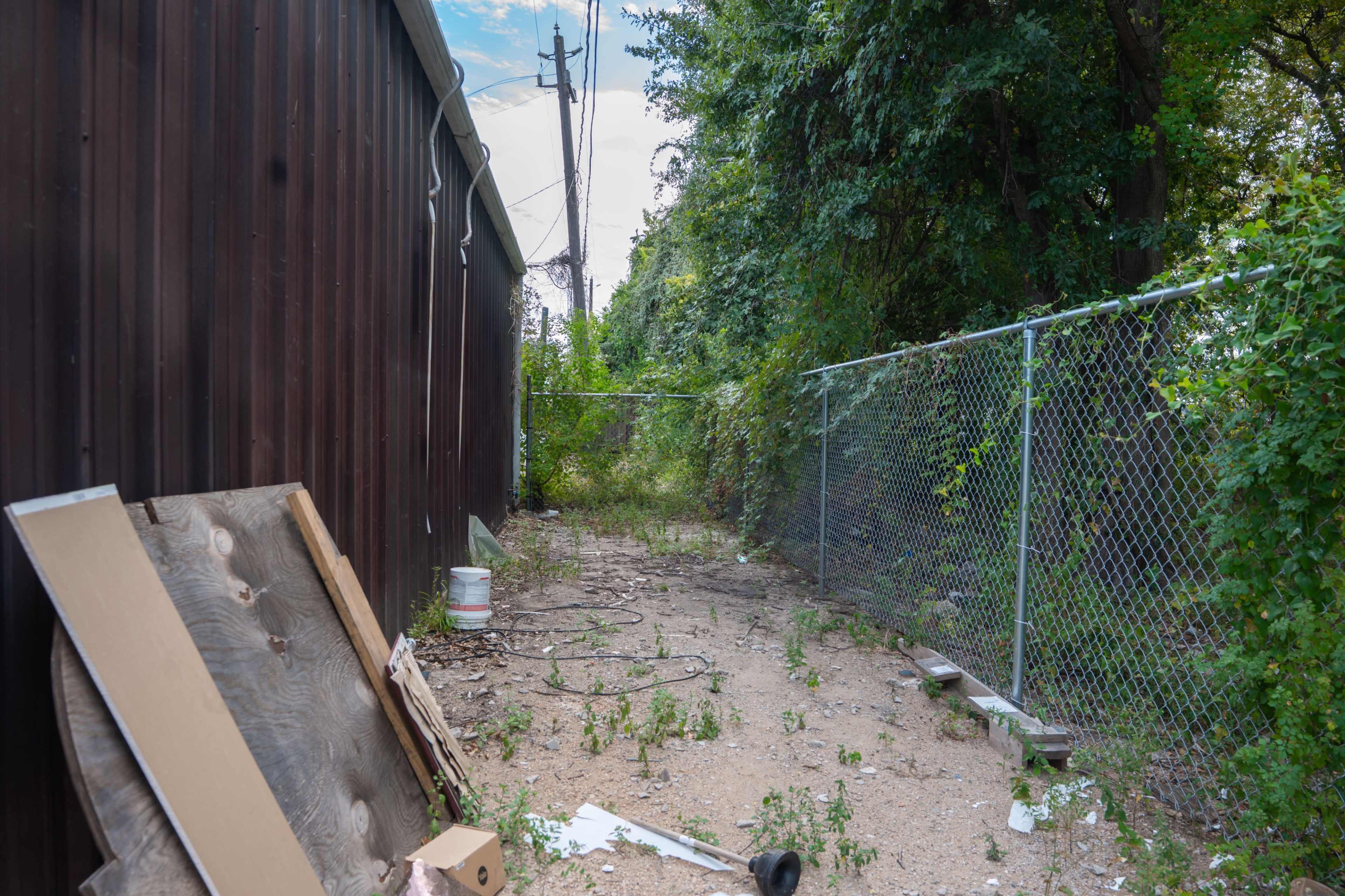 The image shows a narrow, overgrown pathway between a wooden wall and a chain-link fence, littered with debris.