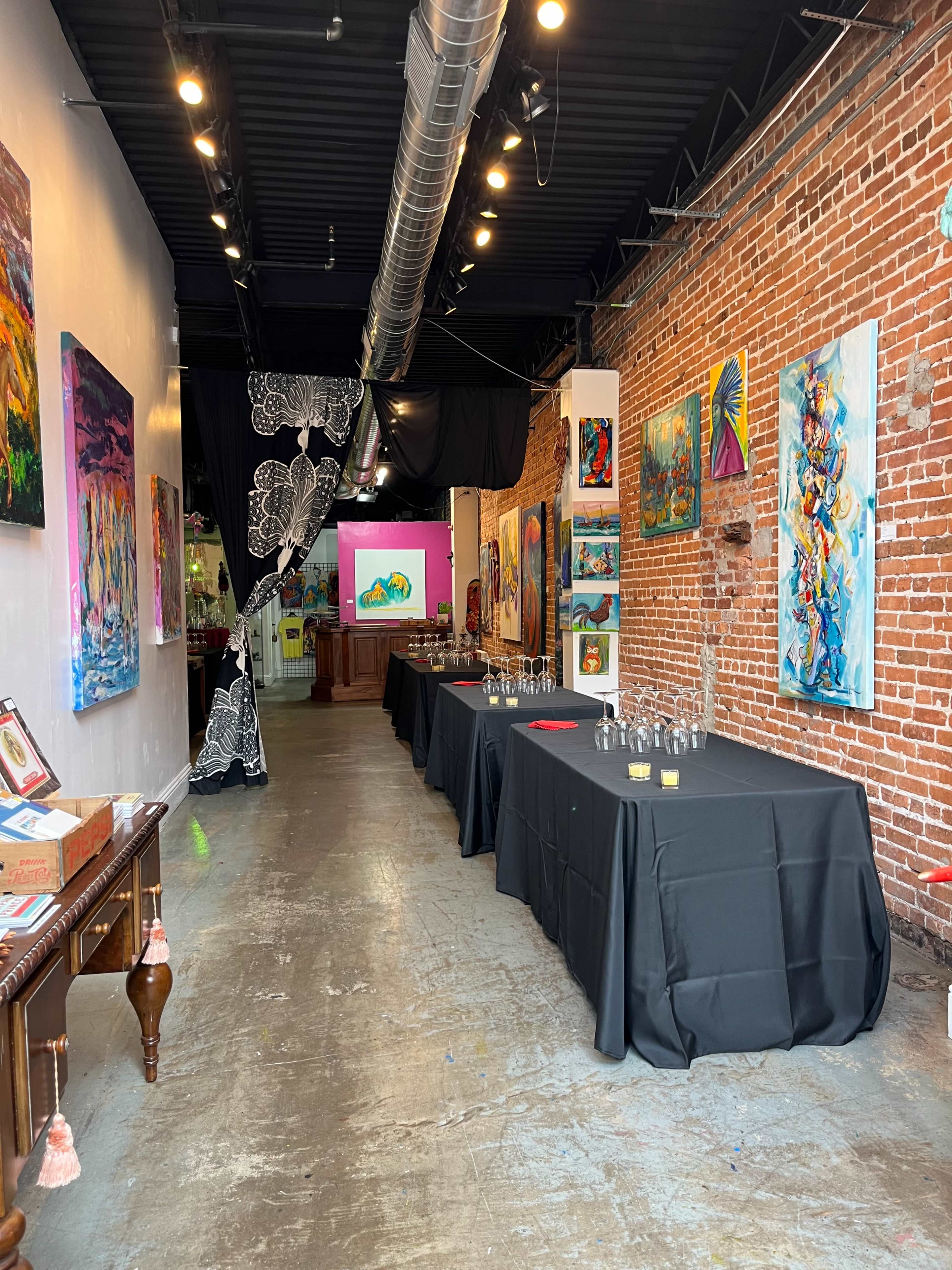 An art gallery featuring brick walls, black tables set for an event, and colorful paintings displayed throughout the space.