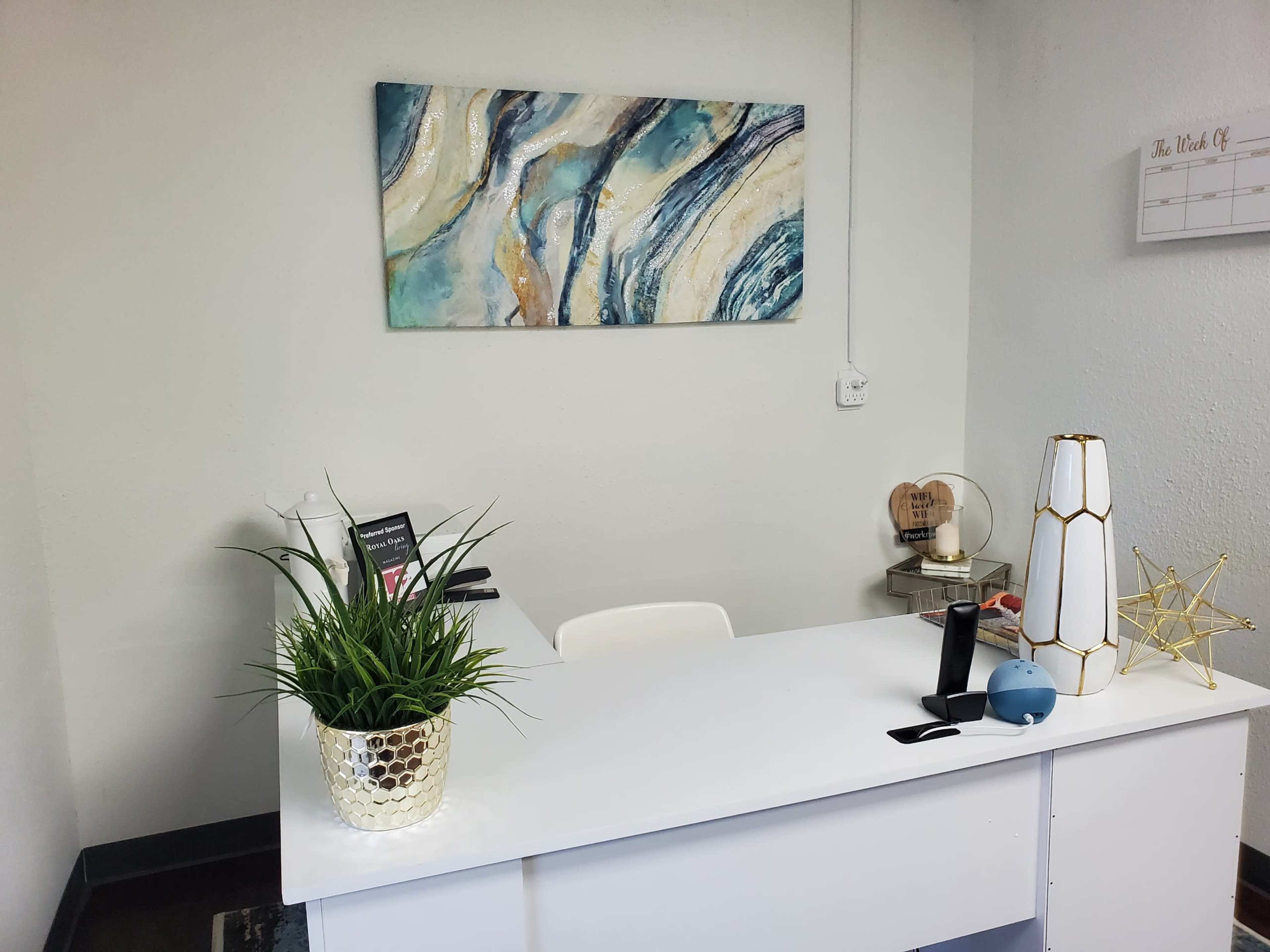 The image shows a modern office reception area with a white desk, a decorative abstract painting on the wall, and a potted plant.