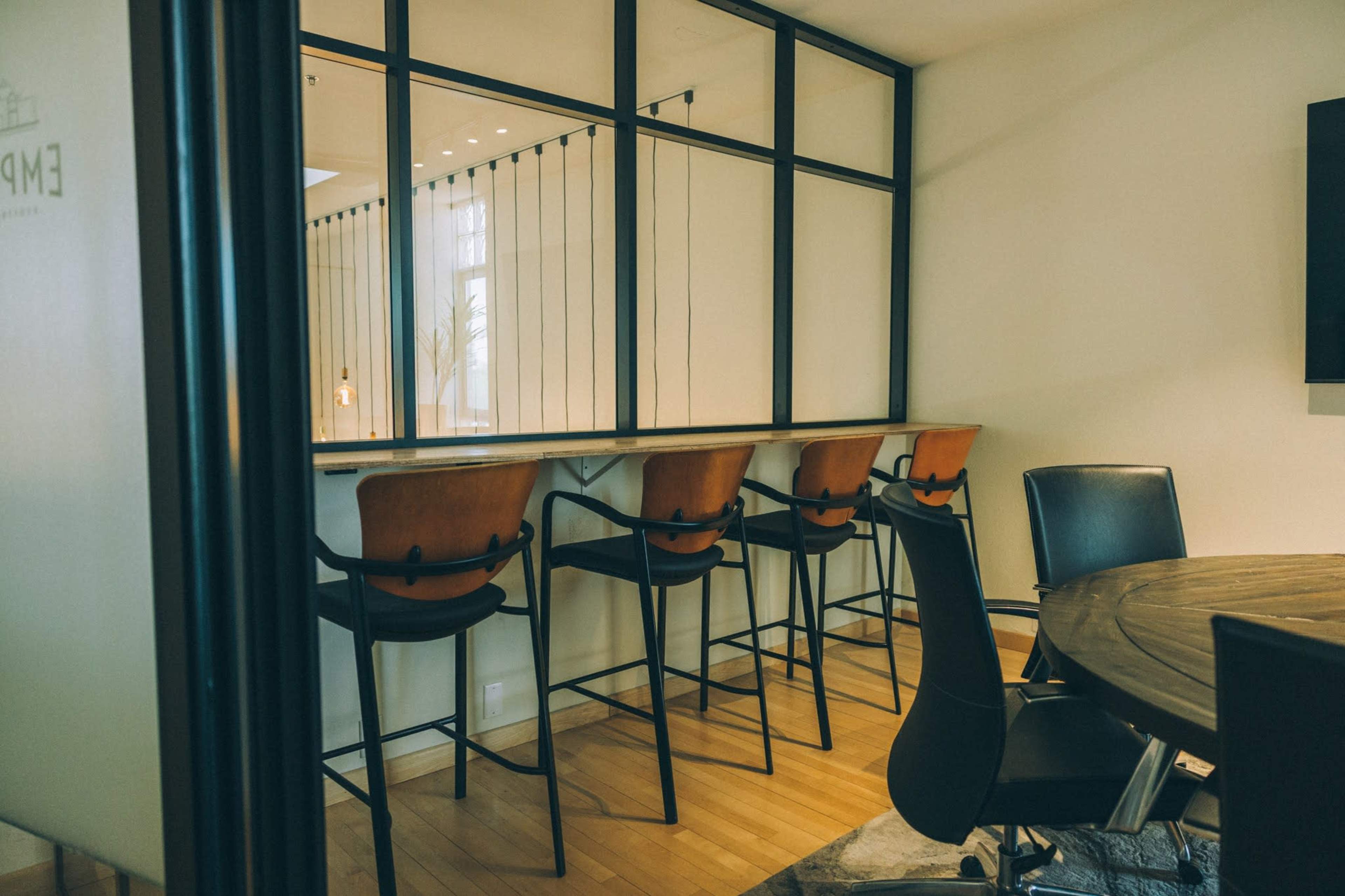 The image shows a modern workspace featuring a row of high-backed chairs along a counter and a circular table in a well-lit room.