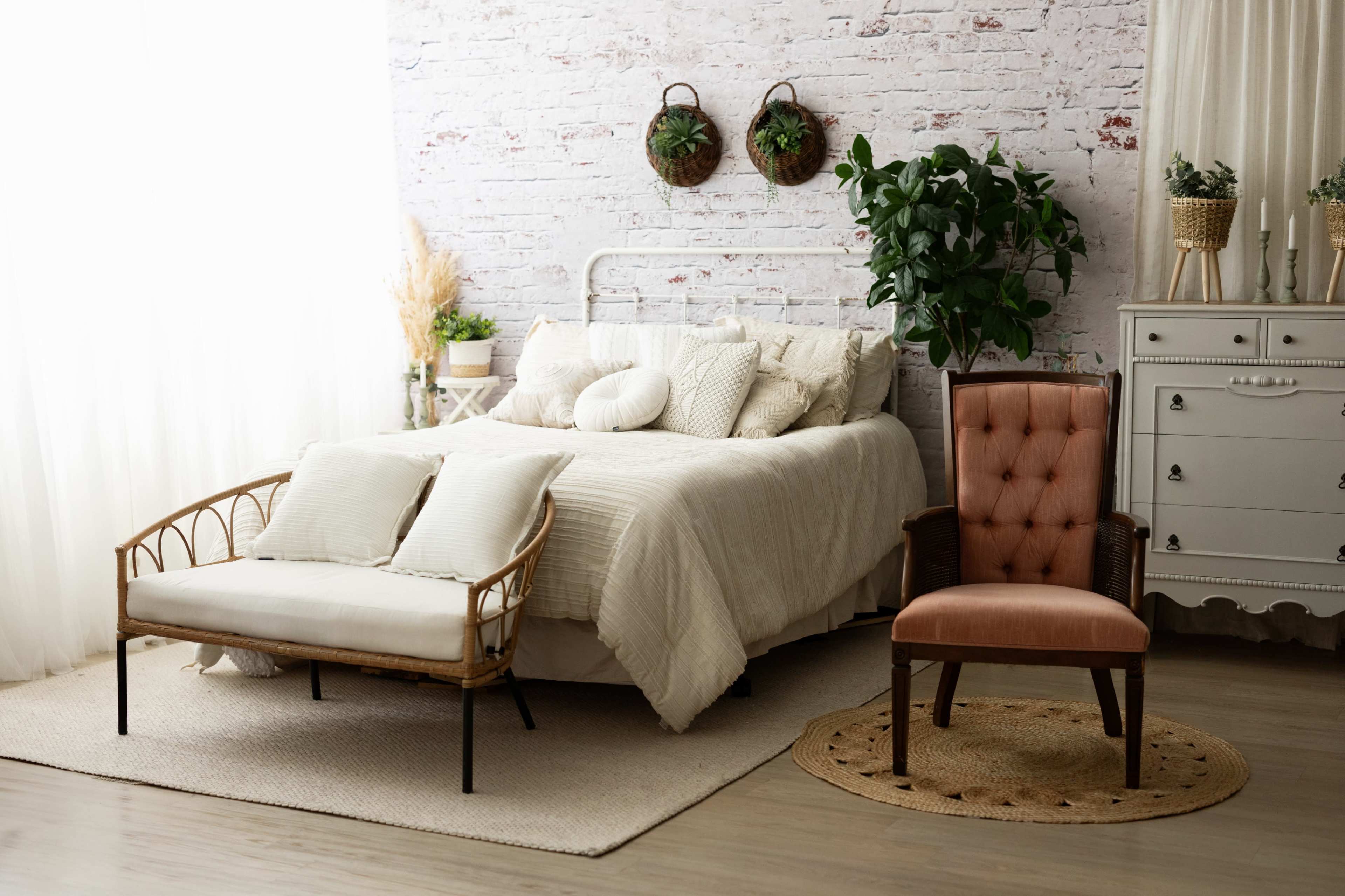 A bright bedroom features a white bed with textured pillows, a rattan bench, an upholstered chair, and greenery against a brick wall.