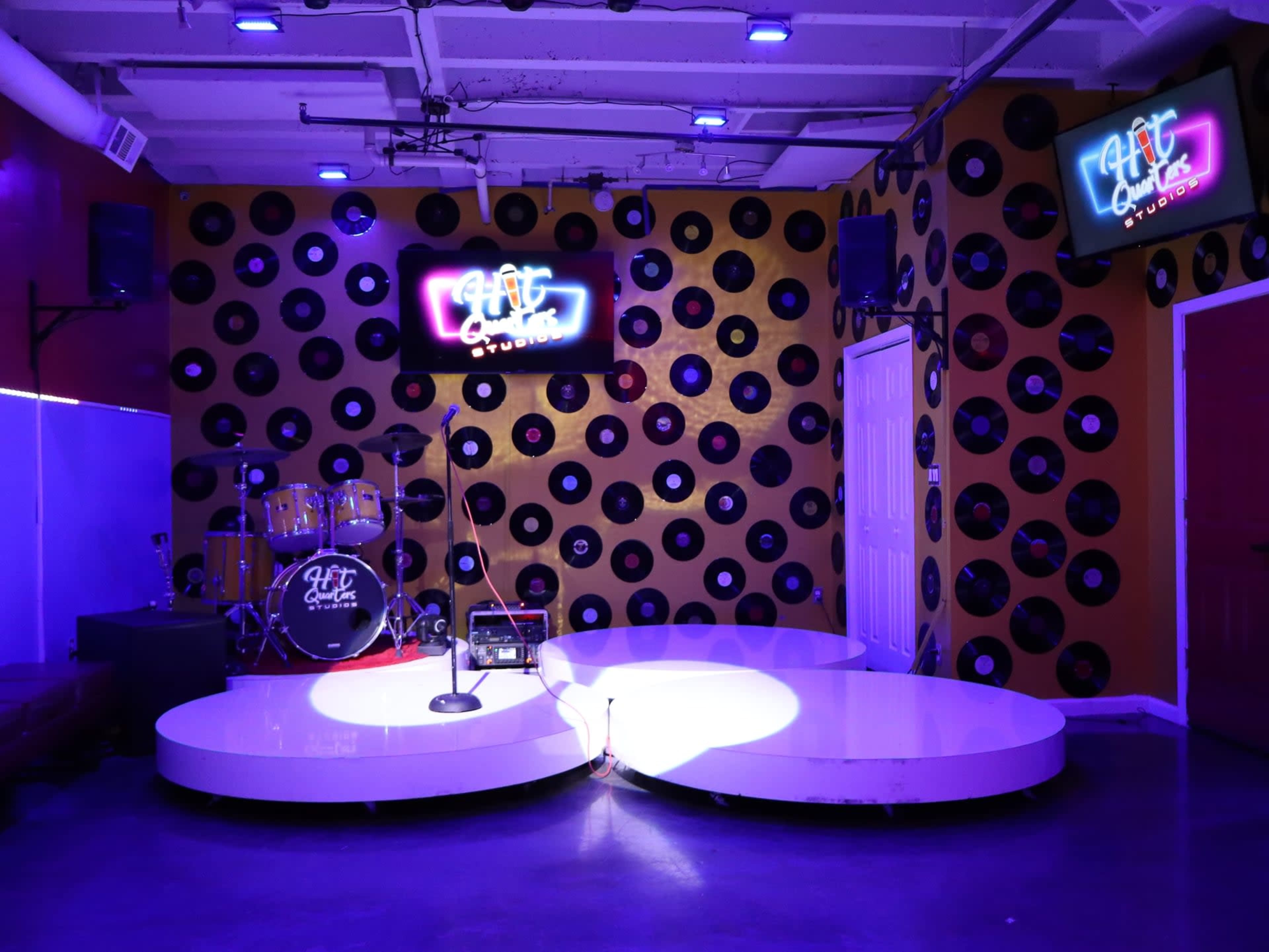 The image shows a stage area featuring a microphone stand, a drum set, and wall decor made of black vinyl records, illuminated by purple lighting.