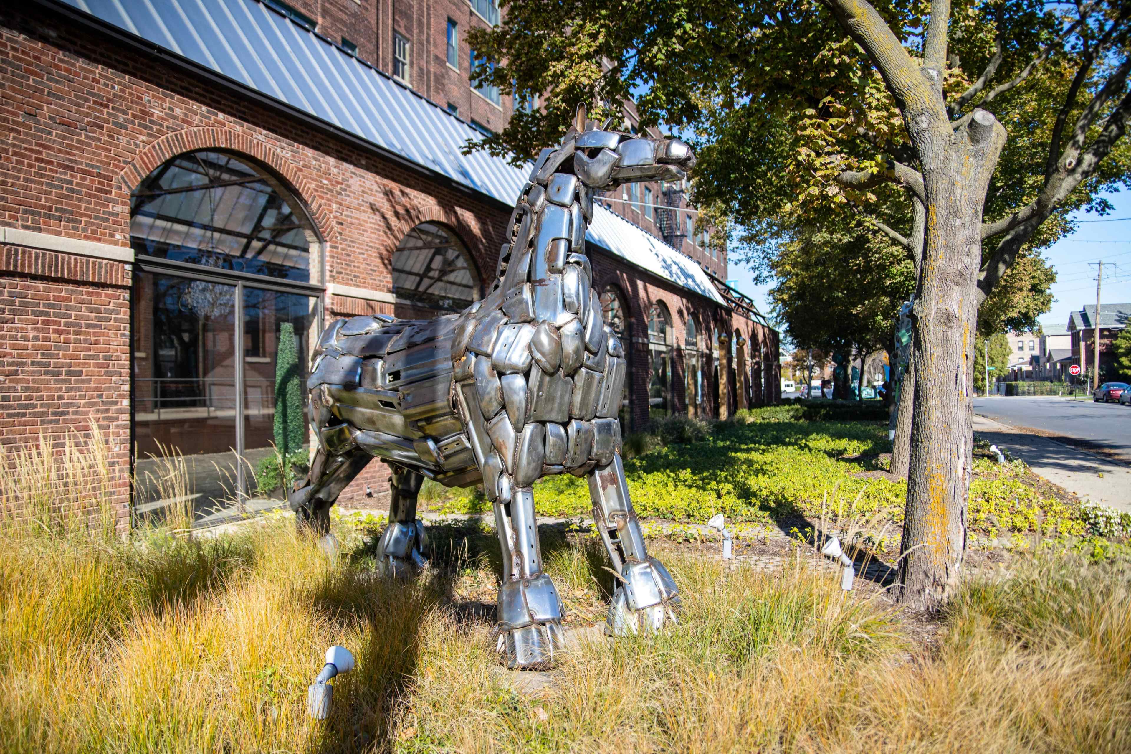 A metallic horse sculpture stands in a grassy area next to a brick building.