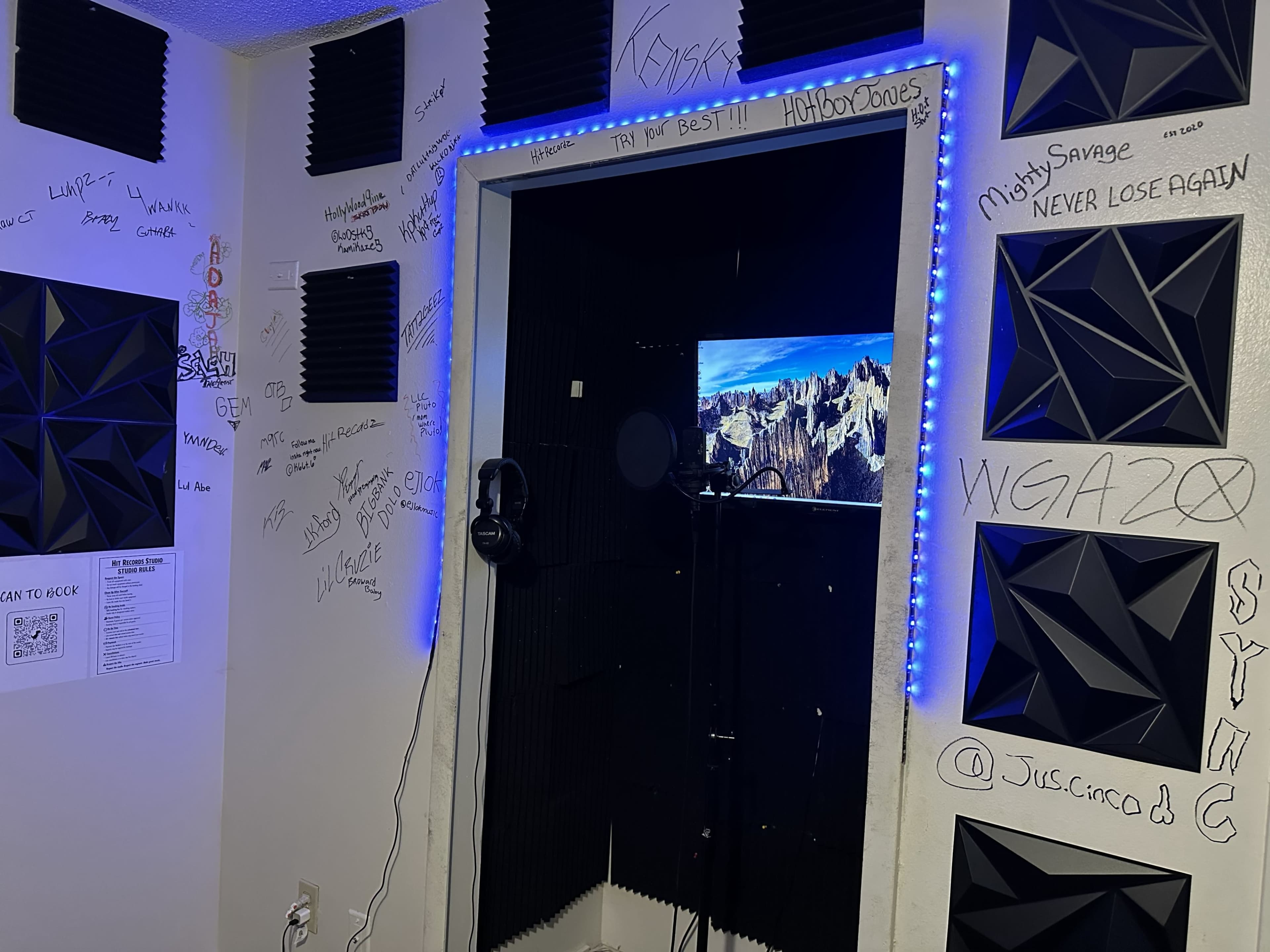 A small recording booth is equipped with soundproofing panels, a microphone, and a screen displaying a mountainous landscape, with inscriptions written on the walls.